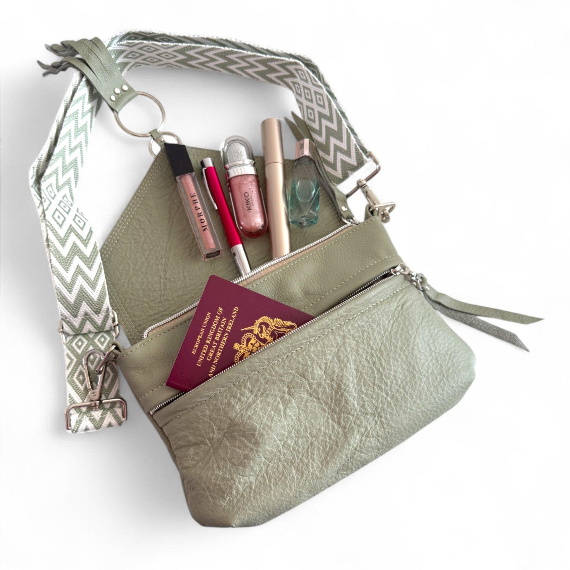 Pale green handbag with cosmetic items spilling out of the main compartment and a passport, peeking out of the front zipper pocket. The Olivia bag is lying on a white background and also shows the matching chevron patterned, woven, adjustable body strap