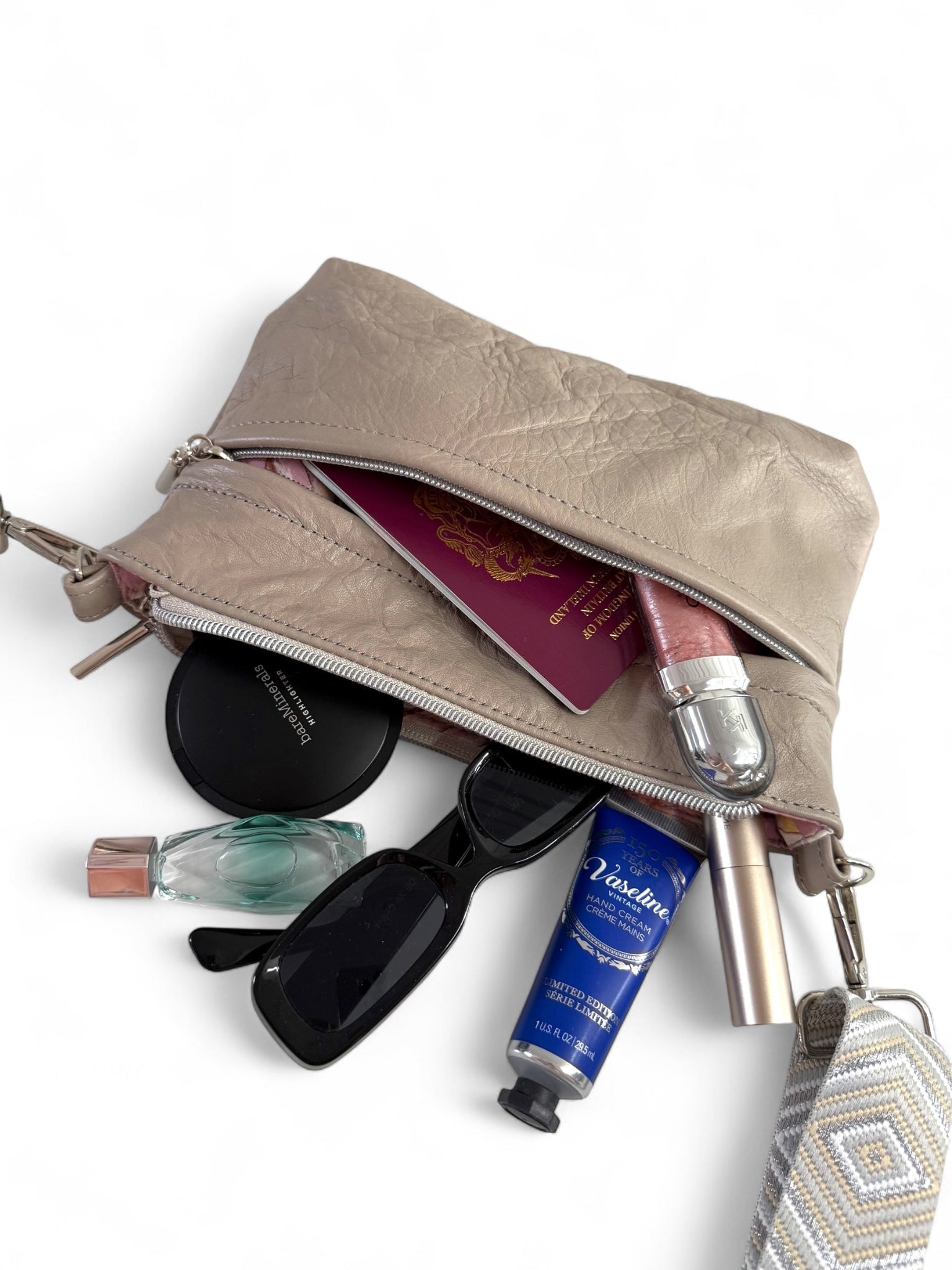 Open grey beige leather bag containing various travel items, spilling contents onto  a white background