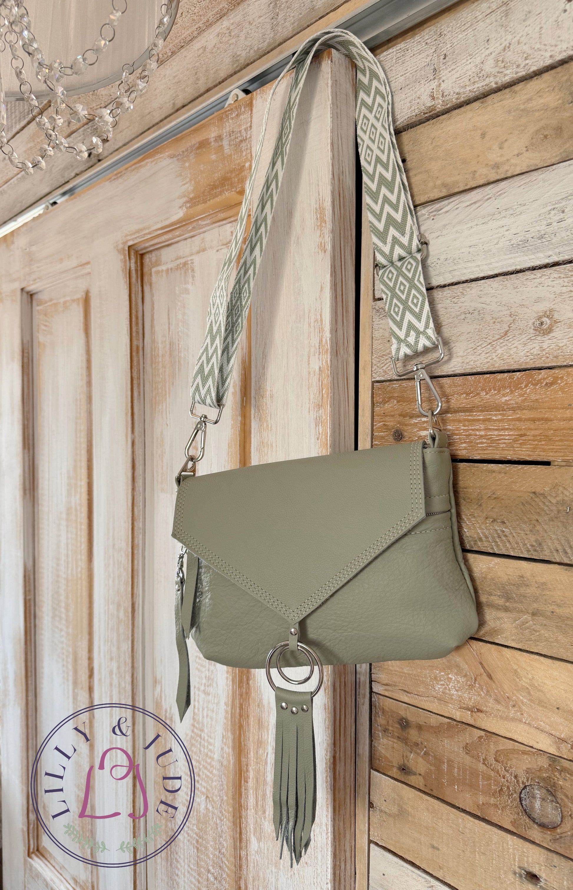 Pale sage green leather crossbody bag, with tassels and matching chevron patterned adjustable strap, hanging on the corner of a rustic looking wooden door. The bag is called Olivia and is handcrafted from rescued leather, in the UK by Lilly & Jude