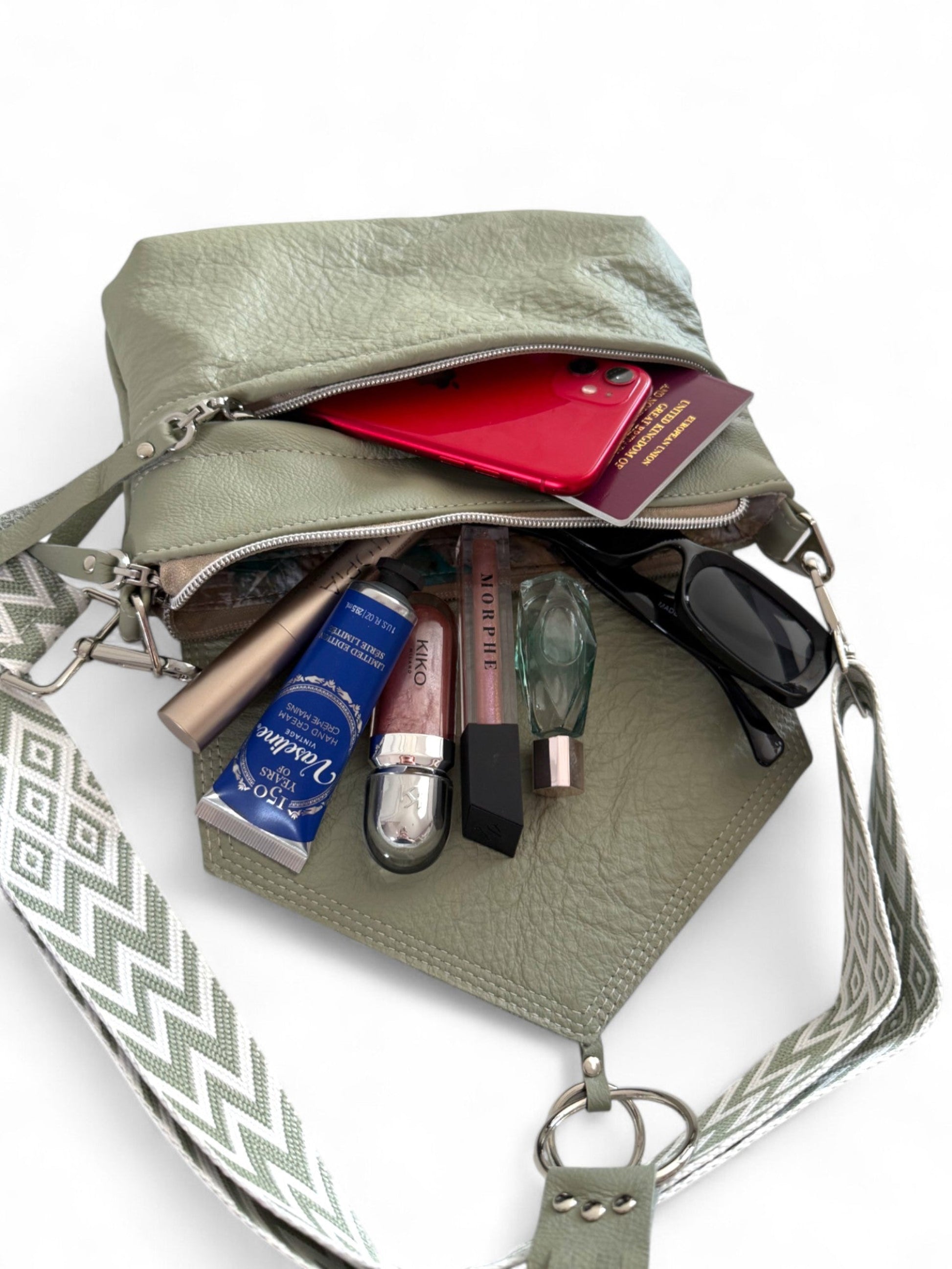 Light olive green handbag with various items including a red phone, sunglasses, and beauty products on a white background. The bag is a handmade, one-off crossbody bag, by Lilly & Jude