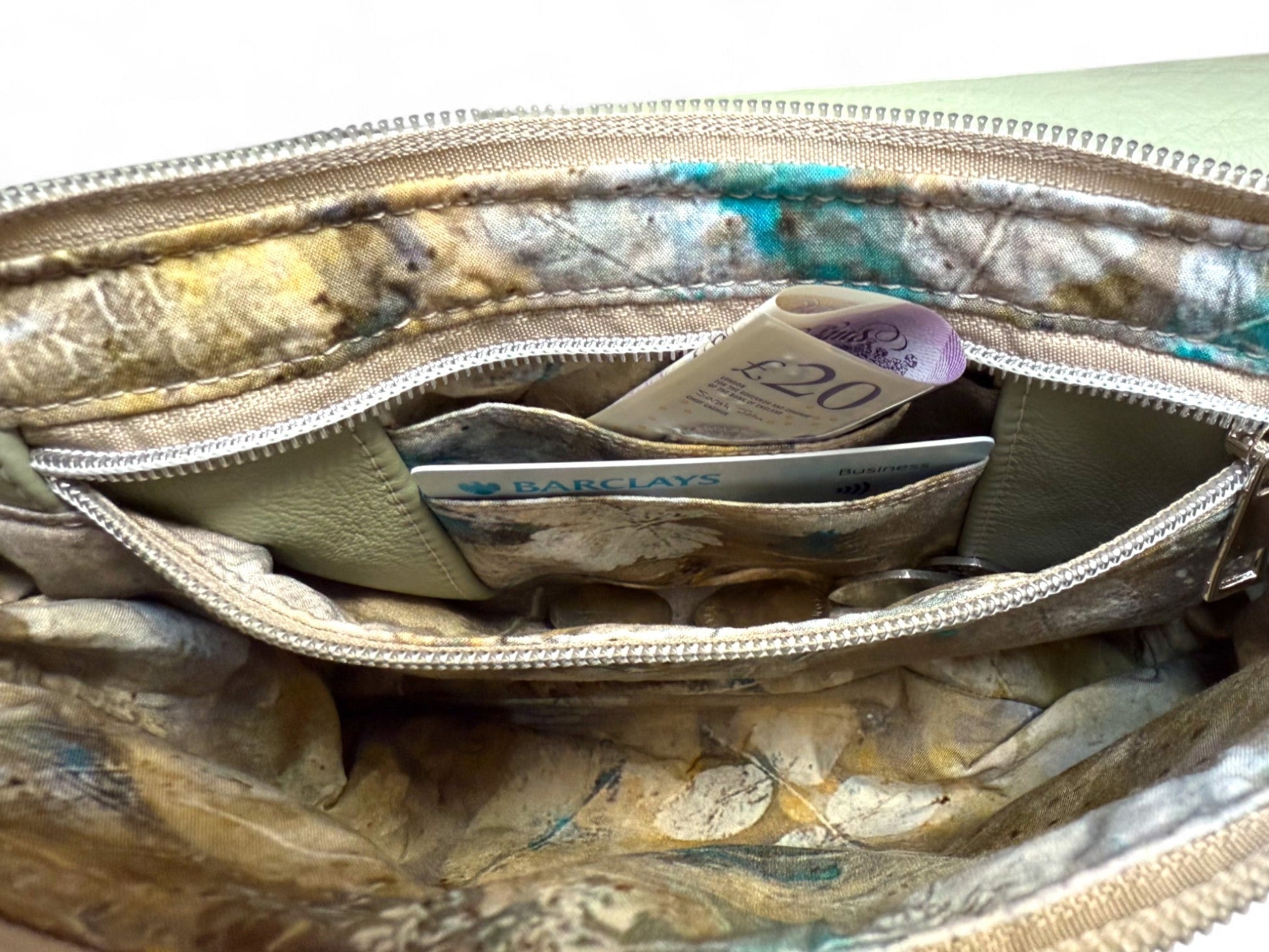 Image shows a pale green Olivia bag. The crossbody bag is open and is showing the inside of the interior zippered pocket, which serves as an integrated purse. There is a £20 note in the back section a bank card in the front card pocket and coins in the bottom of the pocket.
