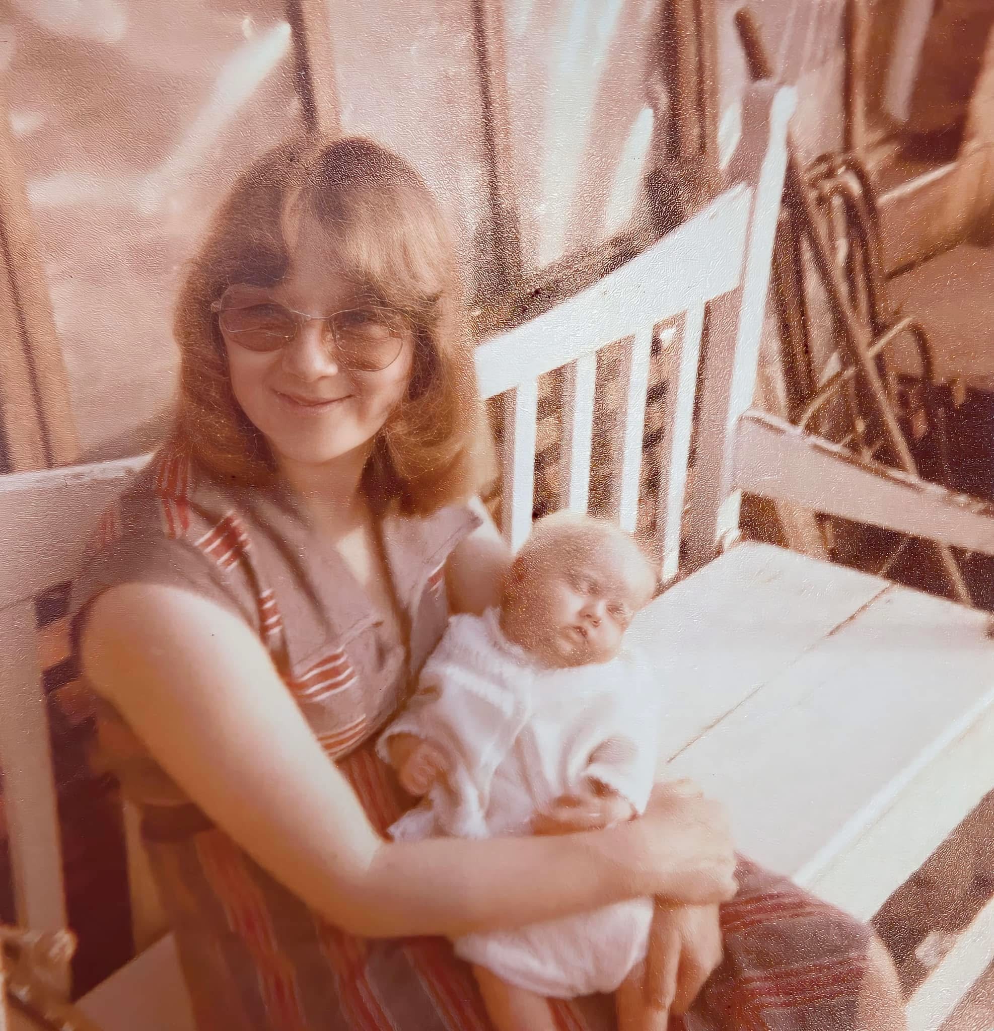 Photo from May 1978 showing Jude, Kerry’s mum, sitting on a white wooden bench in the garden, holding a sleeping newborn Kerry.