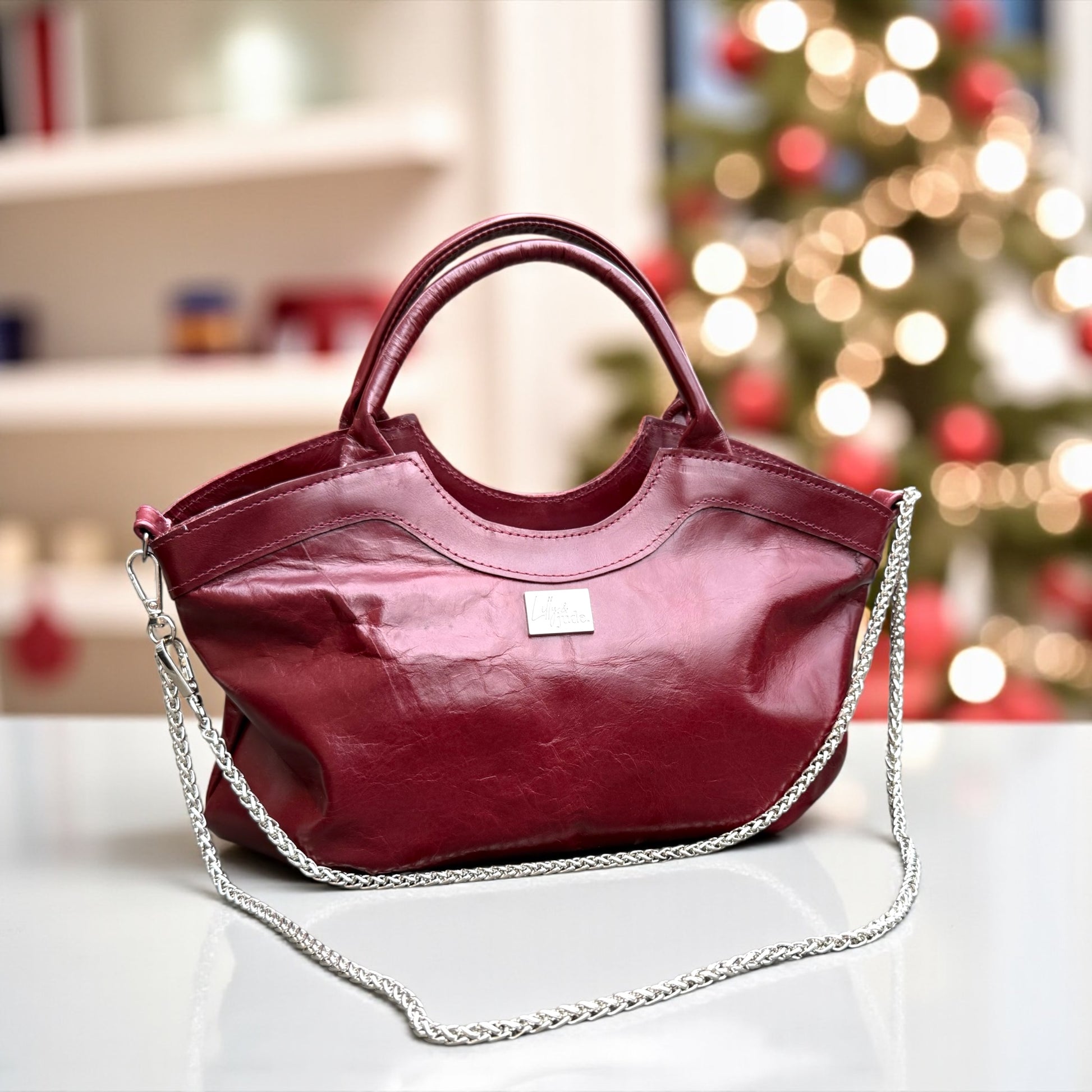 Burgundy leather handbag with a chain detail on a blurred Christmas tree background
