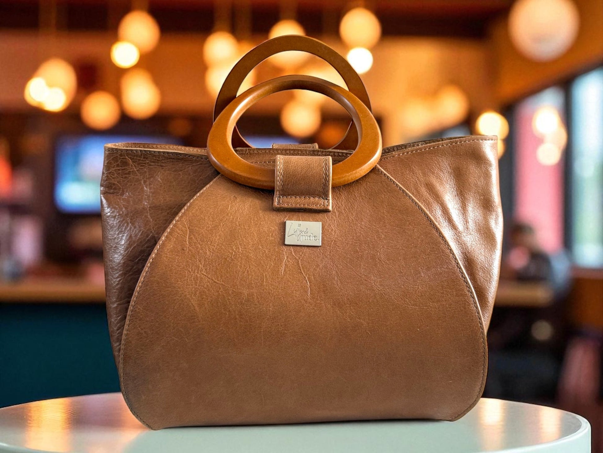 Tan leather handbag with wooden handles on a table in a colourful coffee shop, with bokeh lighting coming from the overhead lighting. It shows the unusual design of the handcrafted bag, which was handmade in the UK by Lilly & Jude