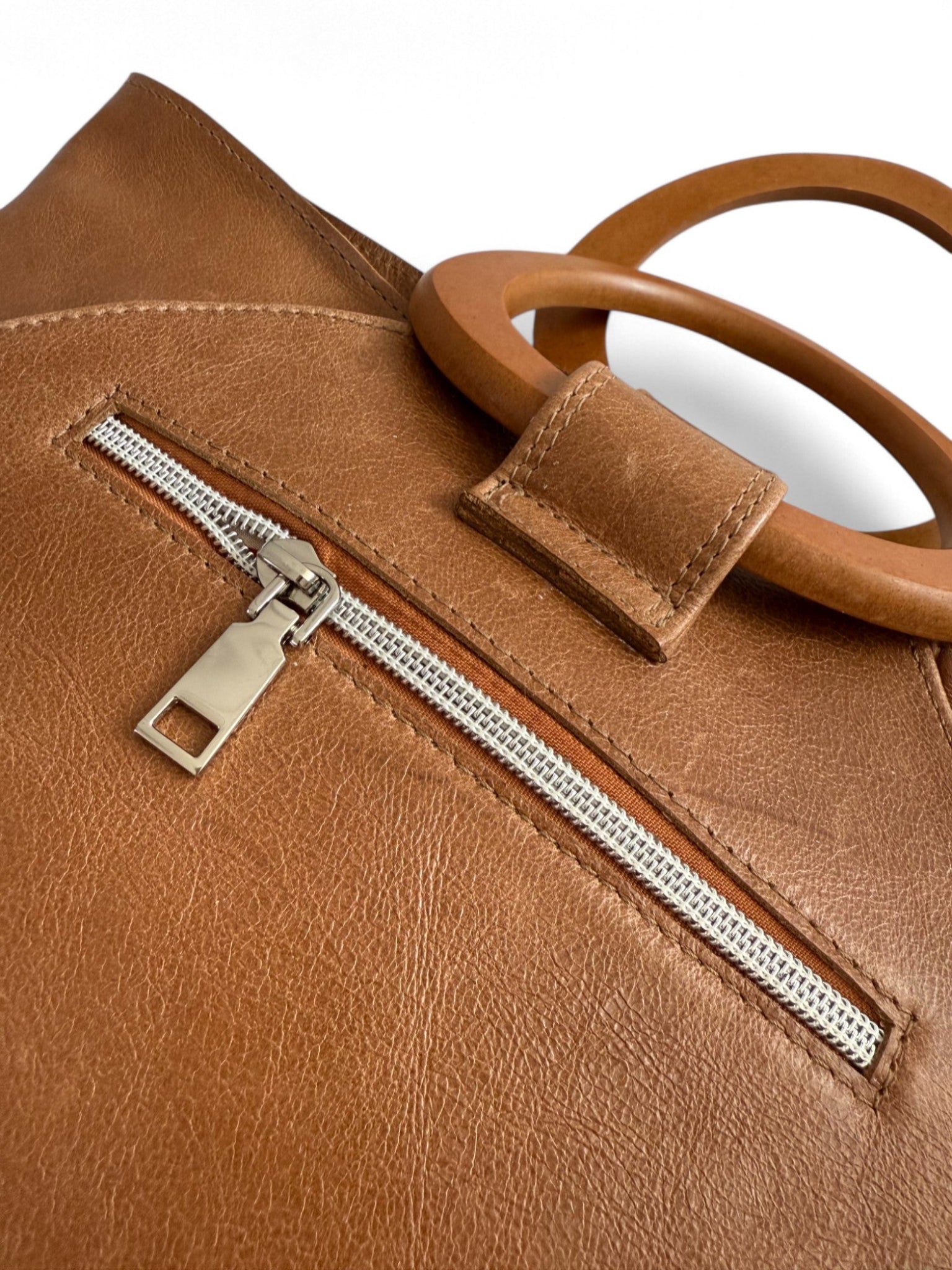 Close-up of a tan leather bag with a zipper pocket and wooden handles on a white background. The image shows the texture and beautiful imperfects in the leather. This bag is called the Tricia and is handmade by Lilly & Jude 