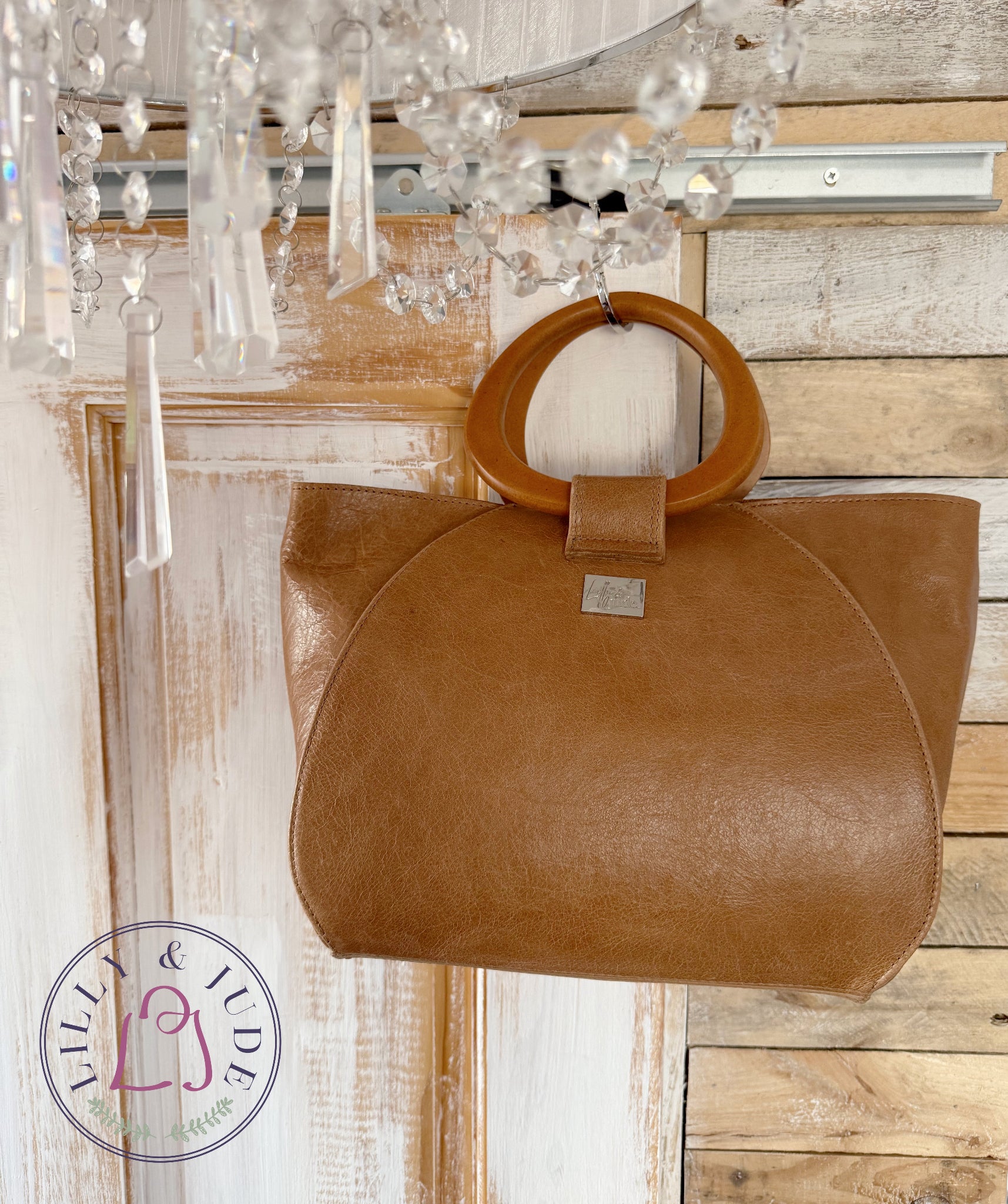 Tan brown leather handbag with wooden handles on a wooden surface with a chandelier in the foreground. The bag is handmade in the UK