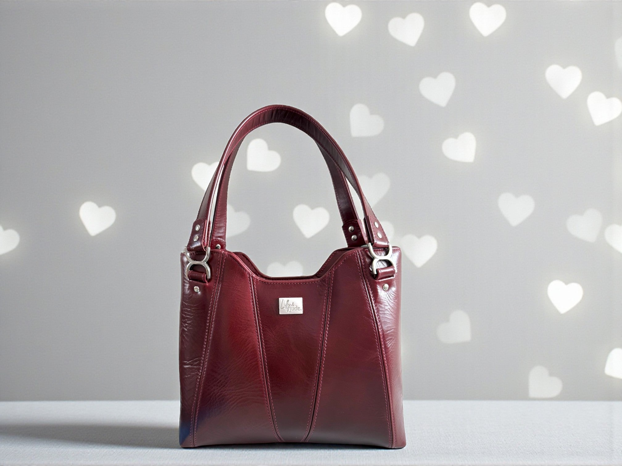 Burgundy red leather handbag with silver hardware and visible Lilly & Jude logo badge. The bag is sat on a light gray background with heart shapes