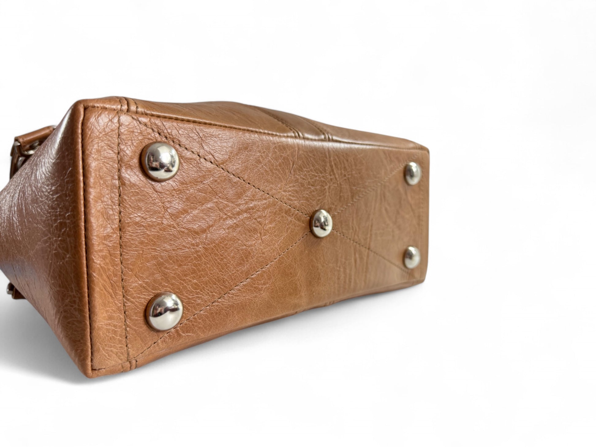 View of the bottom of the tan leather Alaula Handbag. The image shows the bottom of the bag, with decorative stitching and 5 silver bag feet