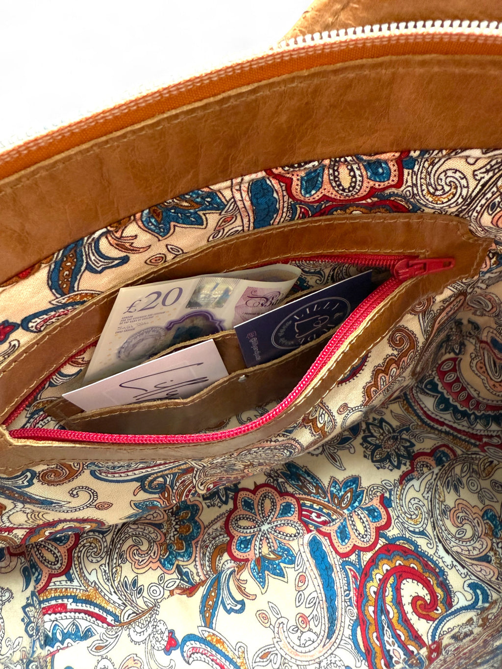 Interior view of the Alaula handbag, showing a closeup of the cream paisley patterned lining and the leather trimmed zipper pocket. Inside the pocket is an integrated card and cash pockets