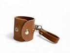 Handcrafted, tan brown leather accessory holder or scarf carrier, with metal accents on a white background. 