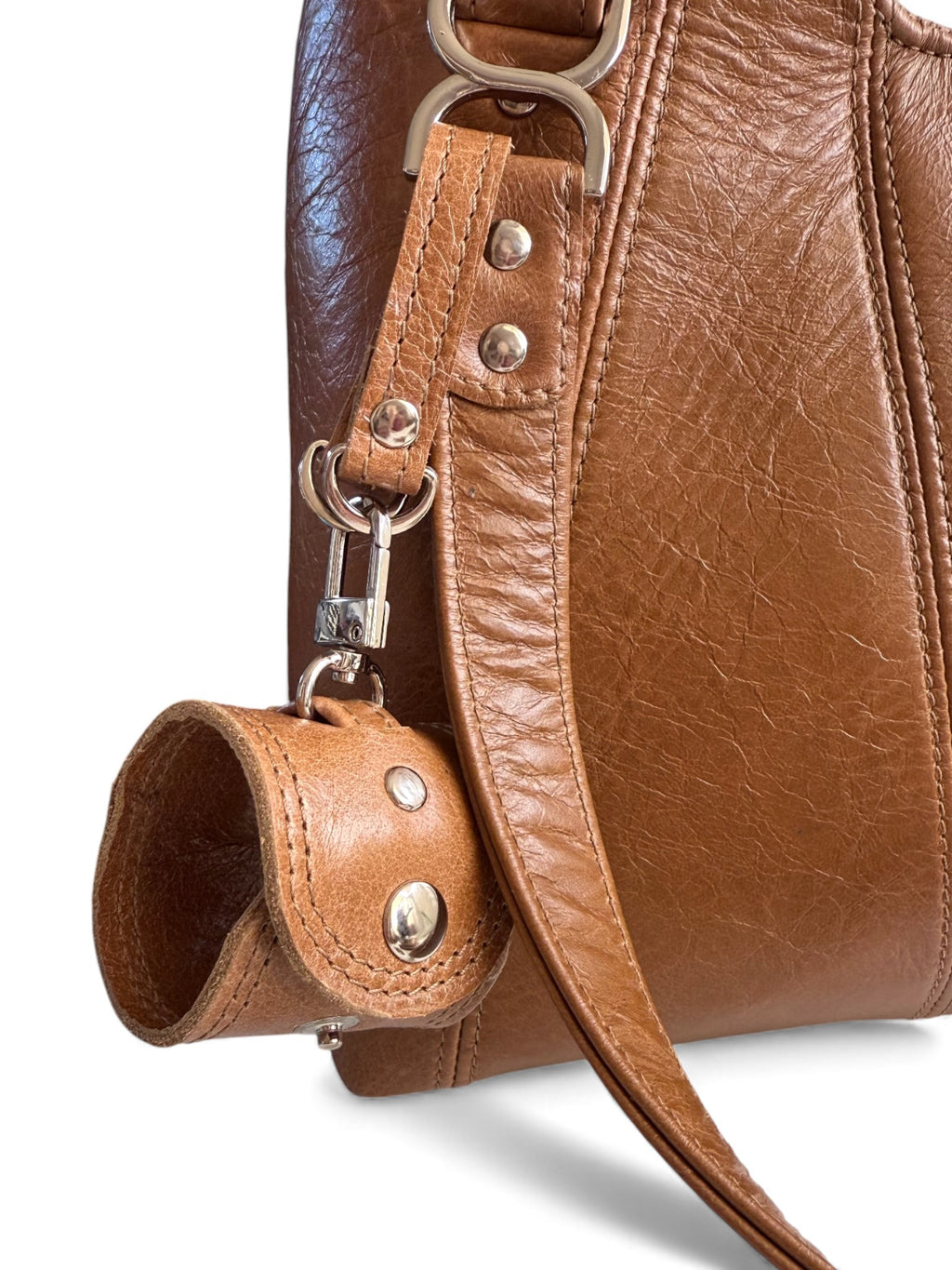 Close-up of a brown leather handbag with a strap and metal hardware on a white background. The strap is designed to hold a scarf or gloves when not in use