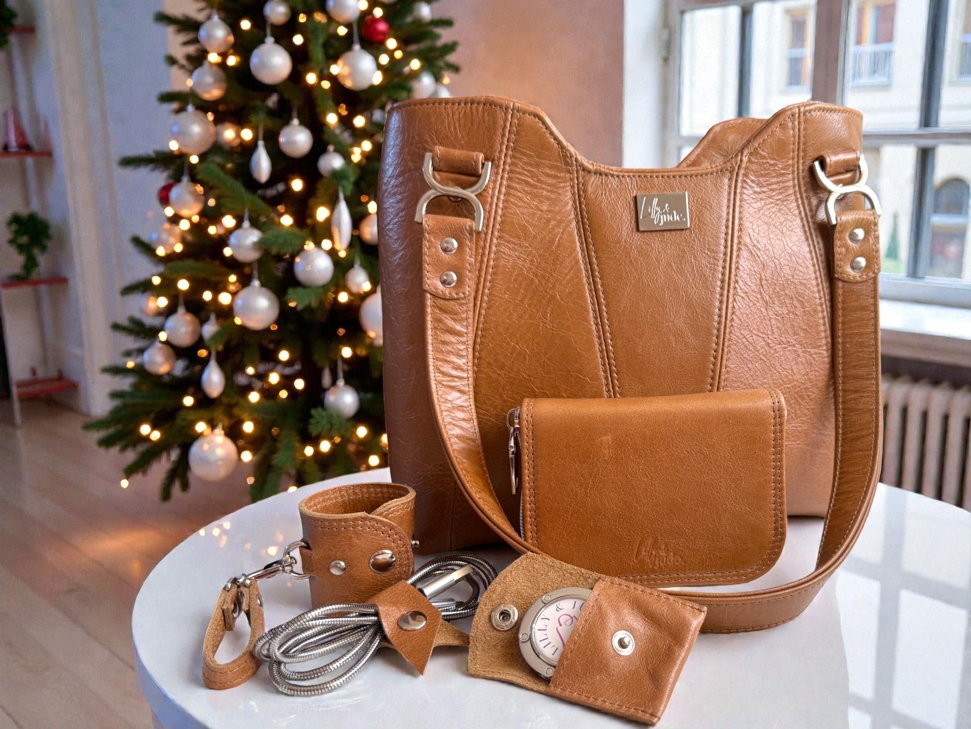 Brown leather handbag with matching wallet / purse, scarf holder, cable keeper and bag hook in a matching pouch, on a table in front of a decorated Christmas tree.