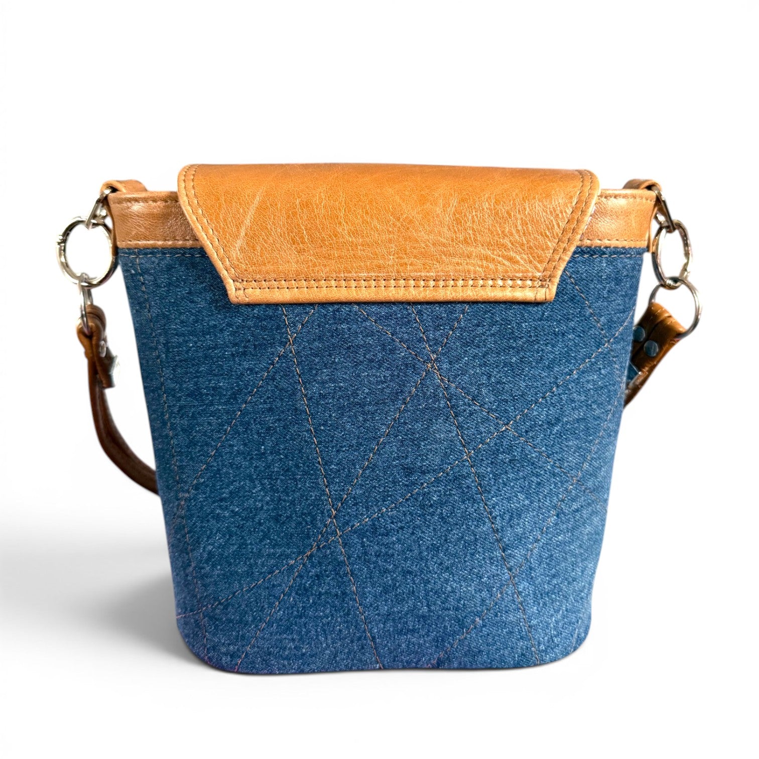 View of the back of the Belle, a blue denim bag with brown leather flap top and shoulder strap, sat on a white background