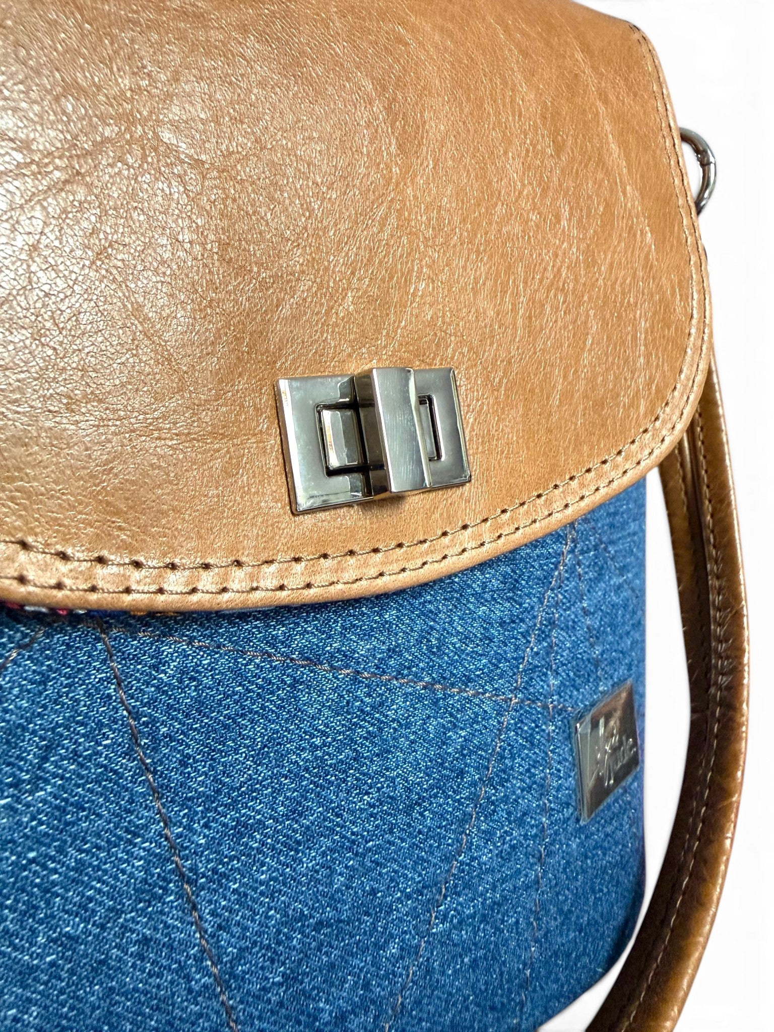 Two-toned handbag with tan leather flap top with silver twist lock and blue denim bottom section on a white background