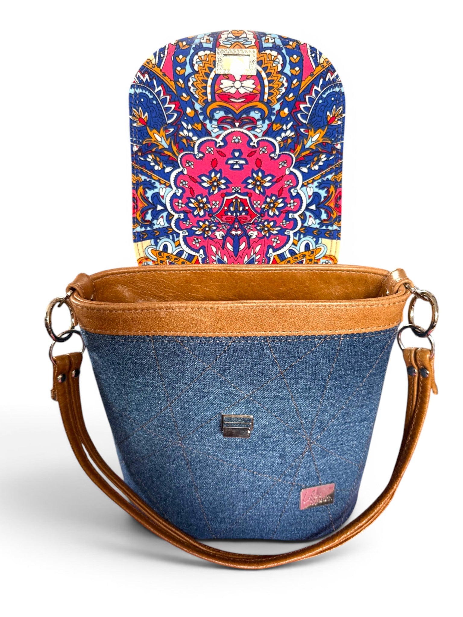 Blue denim bucket bag with tan brown leather strap and trim. The flap is open to show a colourful patterned flap lining. The handbag is sat on a white background