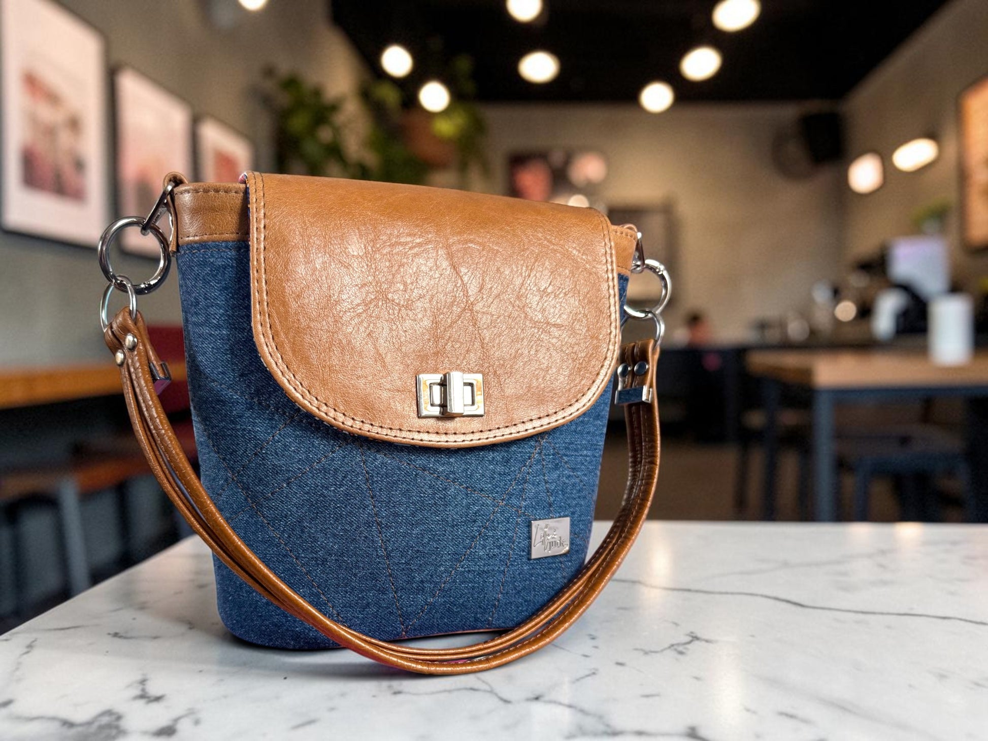 Blue denim and tan brown handbag, with a silver twist lock on the flap, sitting on a marble surface with a blurred cafe background