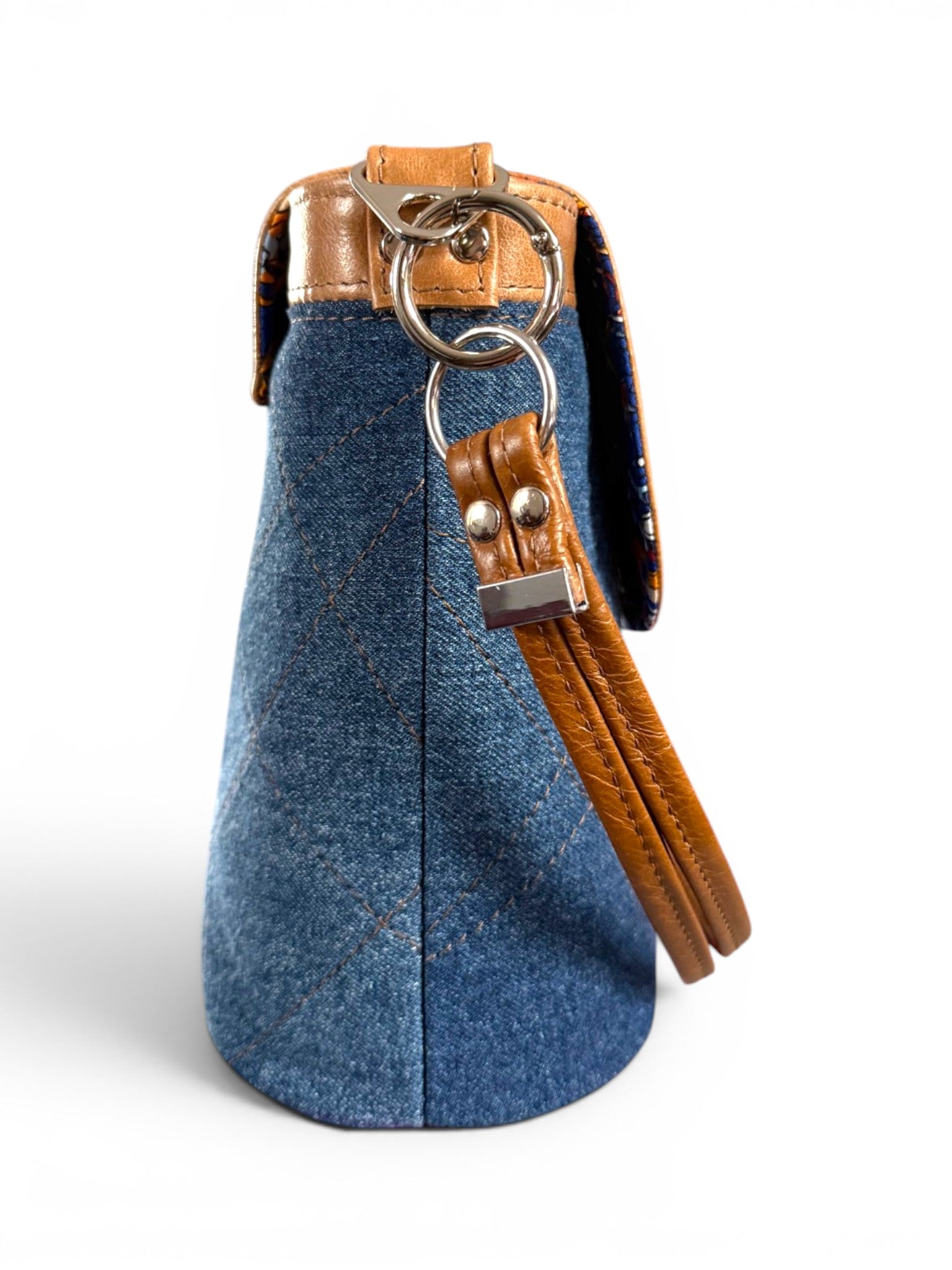Side view of a blue denim bucket style handbag with tan brown leather strap and silver hardware on a white background