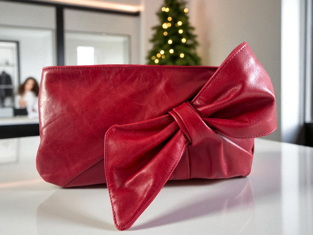 Betty Bow Clutch bag by Lilly & Jude. Red clutch bag with a large bow on a white surface, in a boutique with a lit Christmas tree in the background