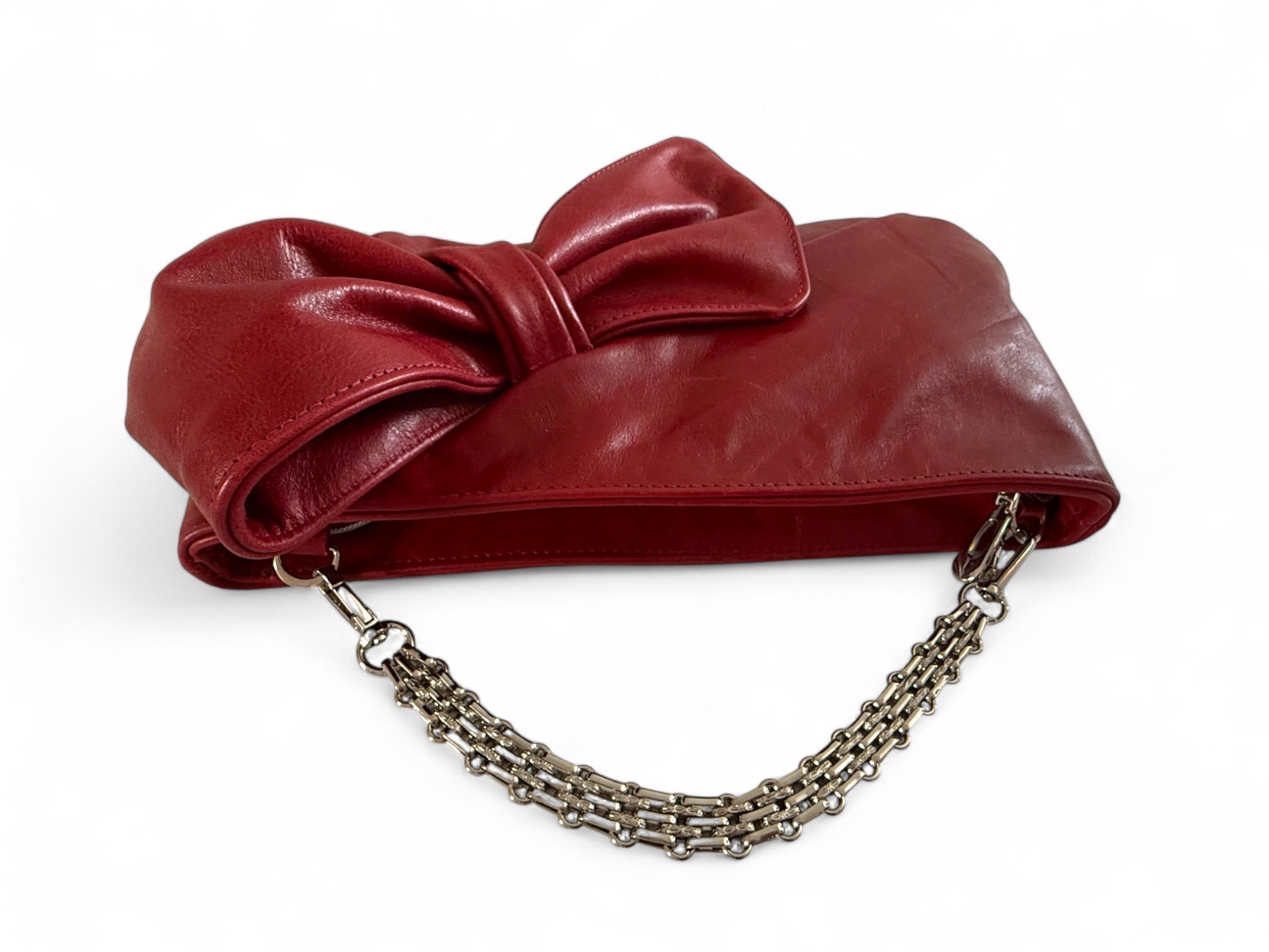 Deep red leather clutch bag with a large bow and chain strap on a white background. The image focusses on the removable silver bracelet style strap. 