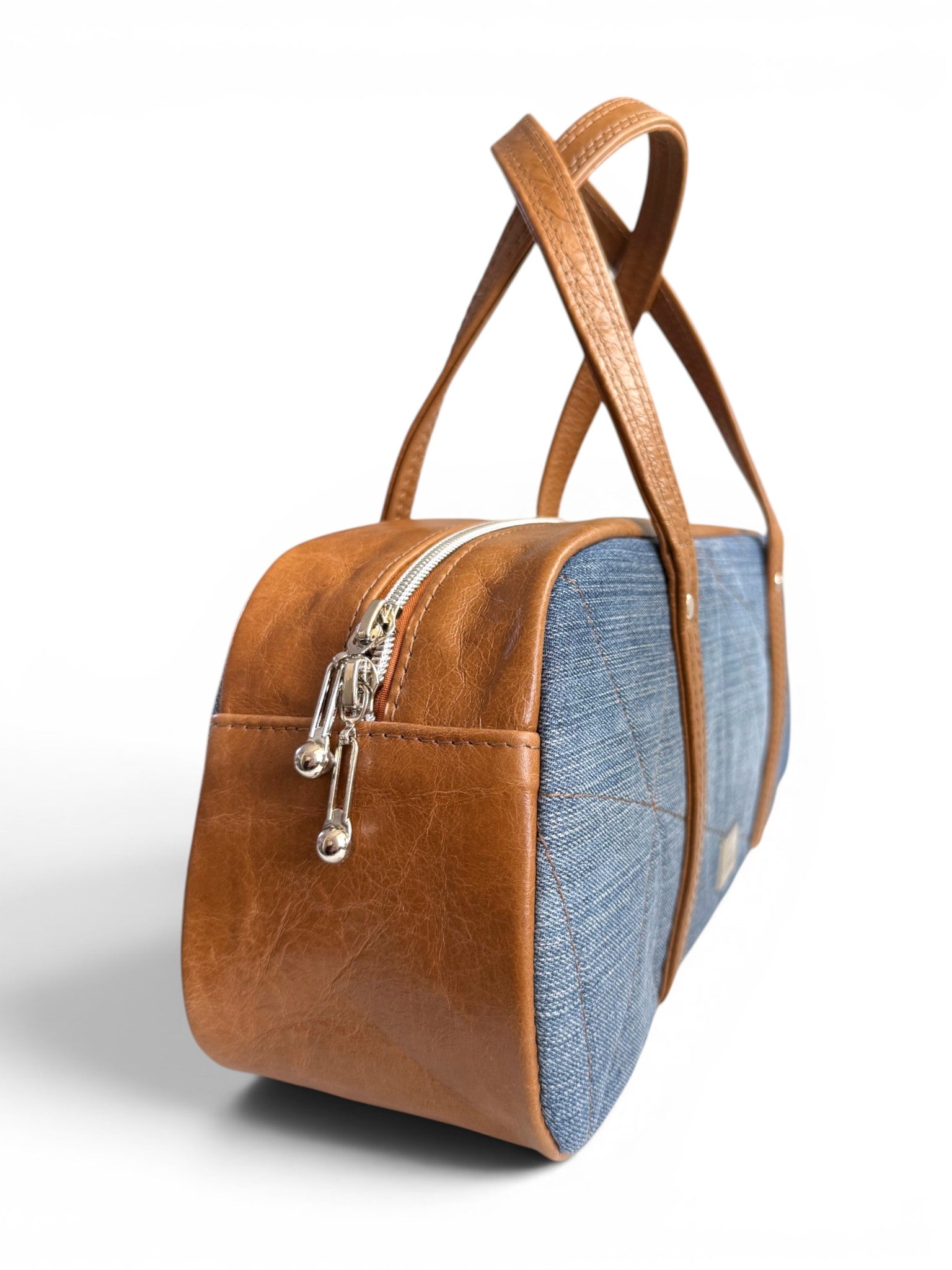 Side quarter view of the Cassie bowling style handbag, highlighting the genuine leather tan gusset, silver zipper pulls and upcycled denim front panel, sat on a white background
