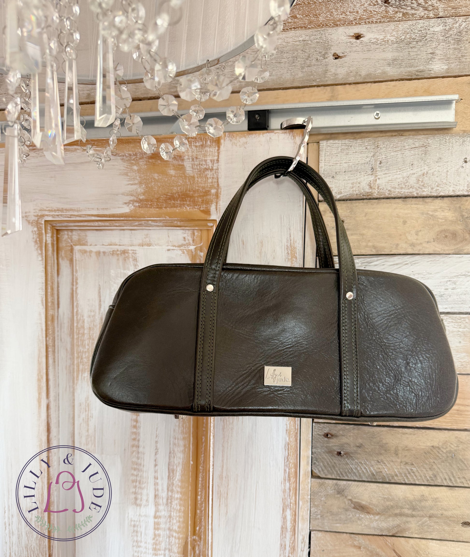 Dark green leather handbag hanging on a wooden door with a chandelier in the background and a Lilly & Jude logo mark 