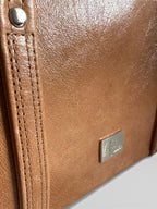 Close-up of a brown leather handbag with the Lilly and Jude brand logo and stitching detail on the strap, on a light gray background