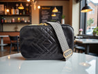 Quilted black leather crossbody bag with chevron patterned woven crossbody strap, sitting on a white table cafe