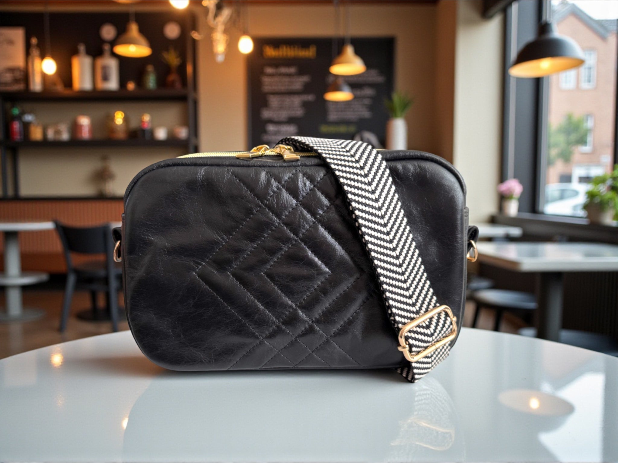 Quilted black leather crossbody bag with chevron patterned woven crossbody strap, sitting on a white table cafe