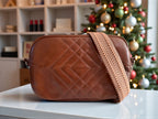Brown quilted leather handbag with a herringbone patterned adjustable, removable strap on a surface in front of a decorated Christmas tree.