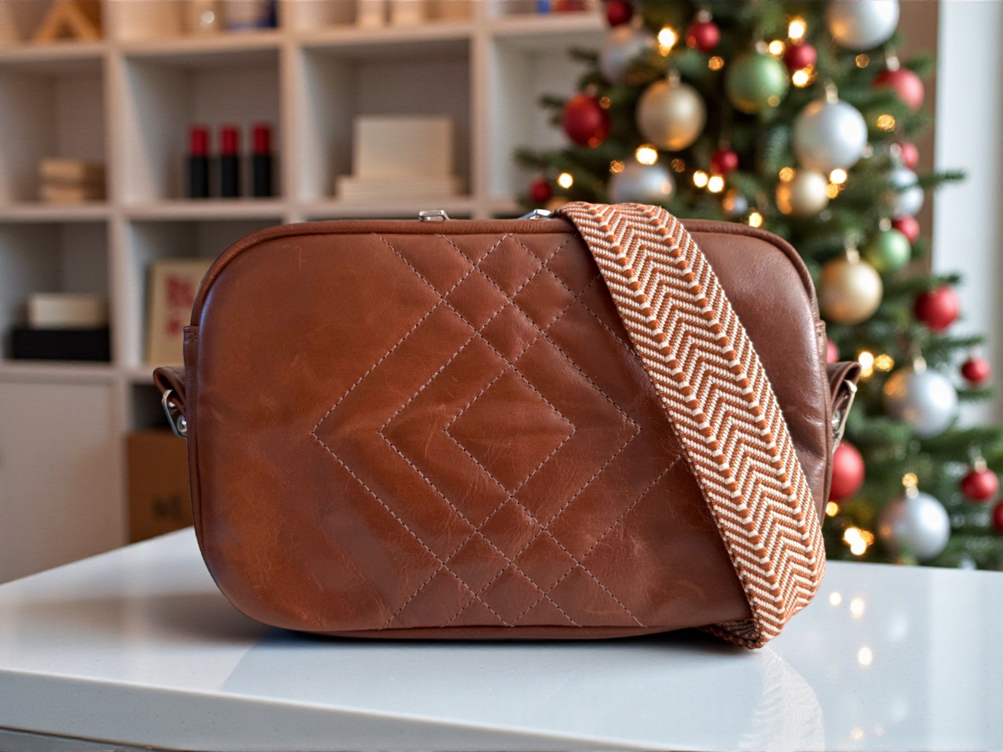 Brown quilted leather handbag with a herringbone patterned adjustable, removable strap on a surface in front of a decorated Christmas tree.