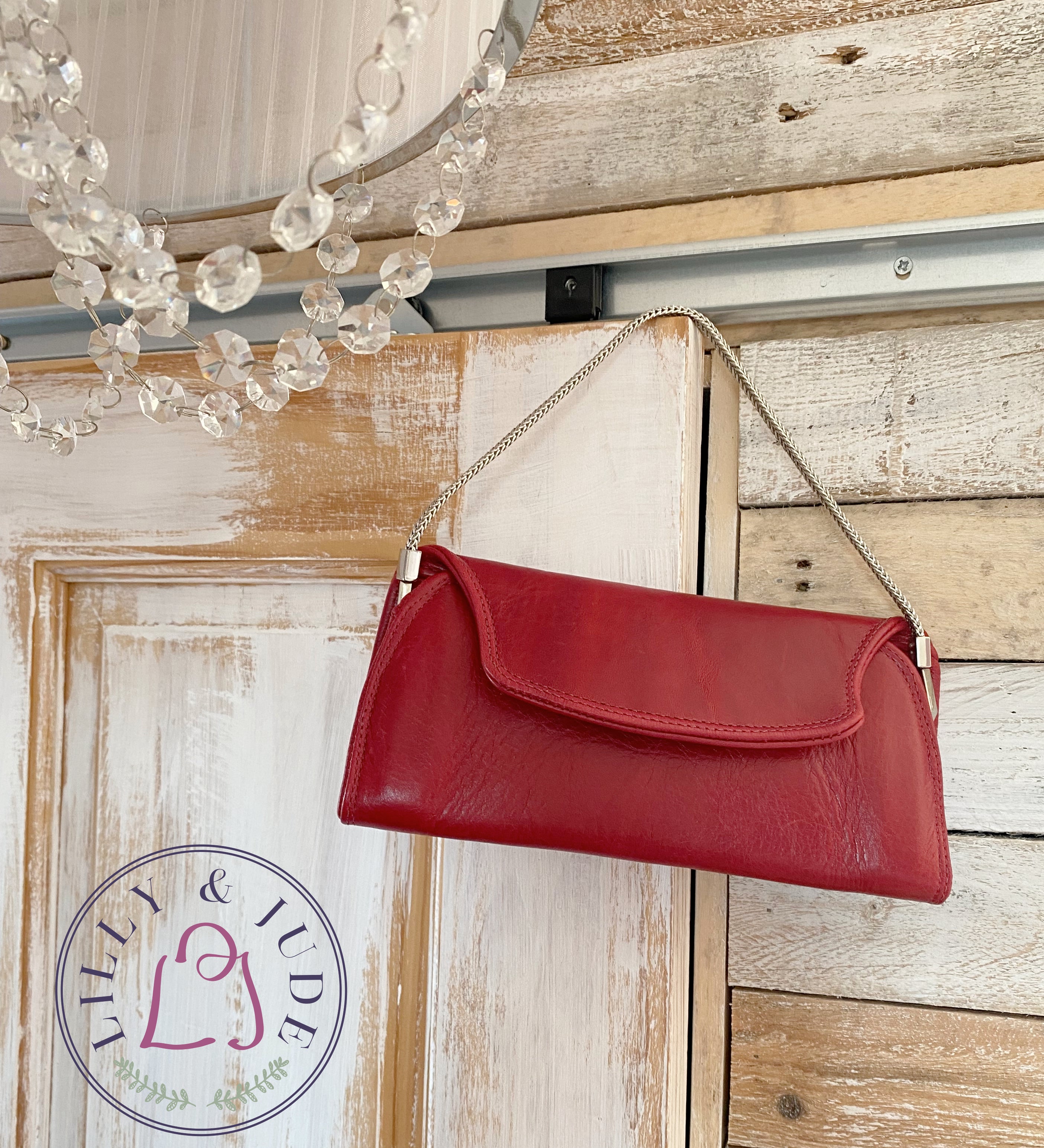 Handcrafted, red leather handbag hanging on a wooden surface with a chandelier in the background and the Lilly & Jude logo in the bottom corner