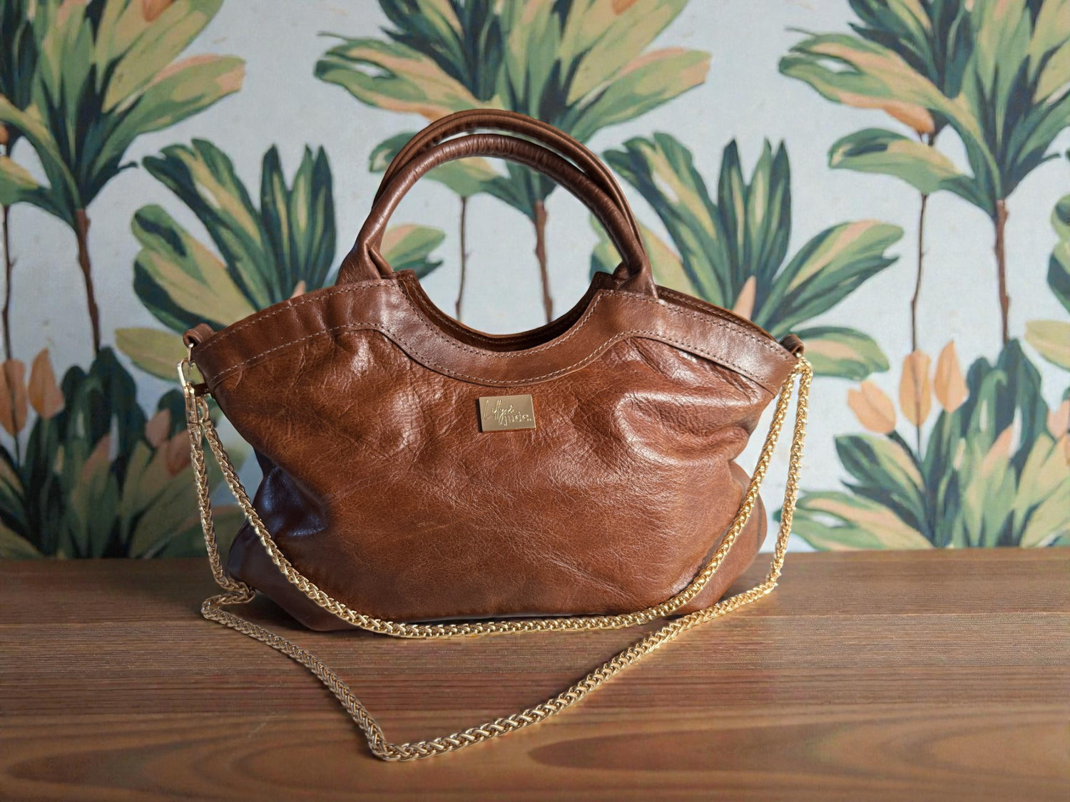 Beautiful, brown leather Flossy handbag, with gold hardware and chain, on a wooden table with leaf patterned wallpaper in the background
