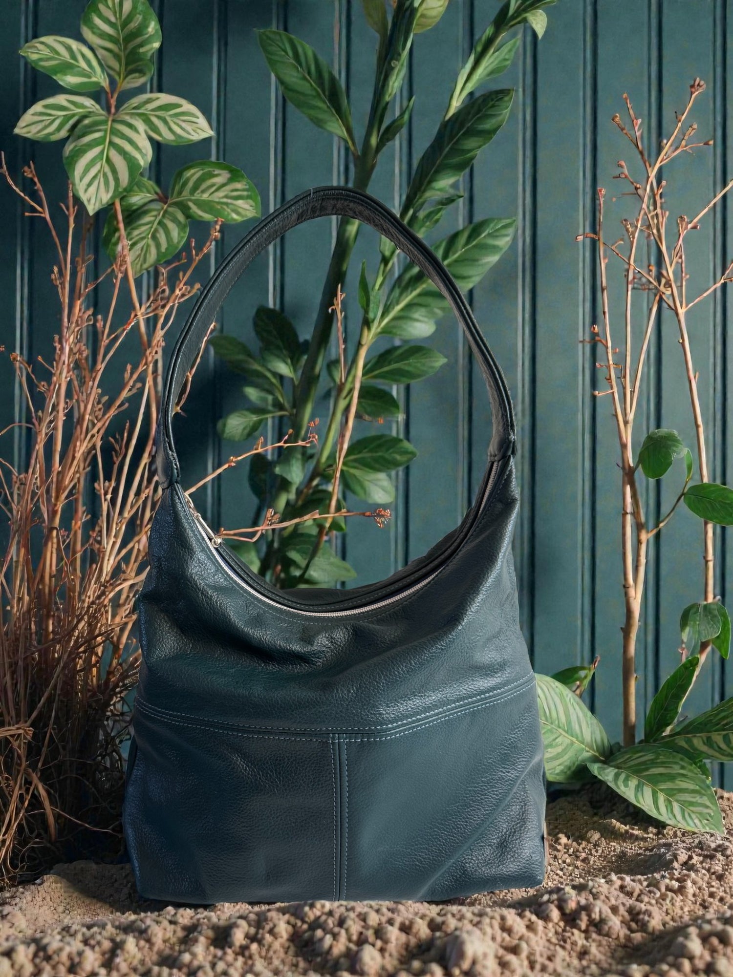 Dark teal coloured leather slouch handbag, sitting in a garden scene, with a wooden fence and plants