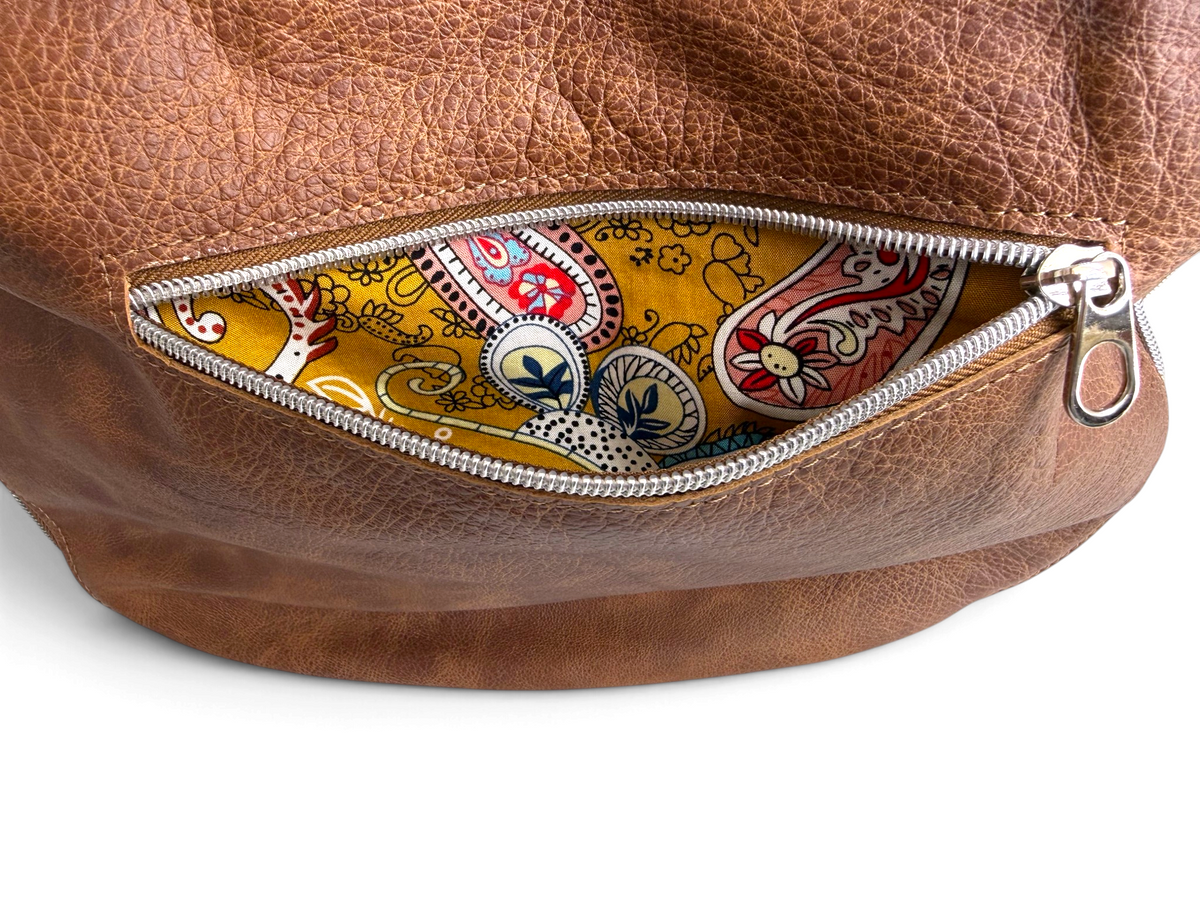 Close-up of a brown leather zippered pocket showing a colorful paisley-patterned fabric lining on a white background.