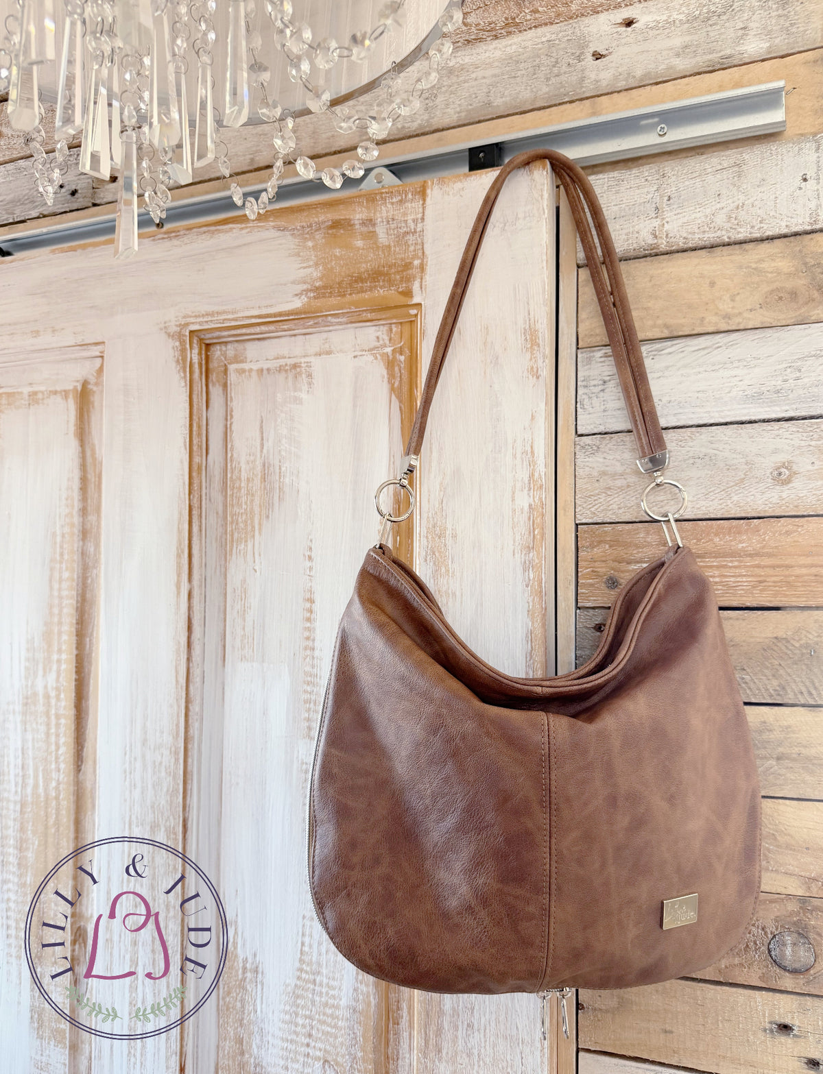 A brown leather slouchy shoulder bag hanging against a rustic wood-panelled wall, with a small Lilly & Jude logo on the lower right corner.