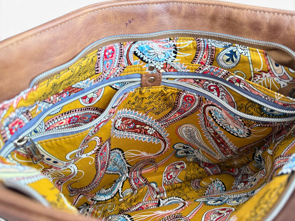 Close-up of the interior of a brown leather bag showing a mustard yellow paisley-patterned lining and 2 slip and zip pockets