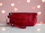 Red leather clutch bag with a wrist strap on a light surface with heart-shaped lights in the background.
