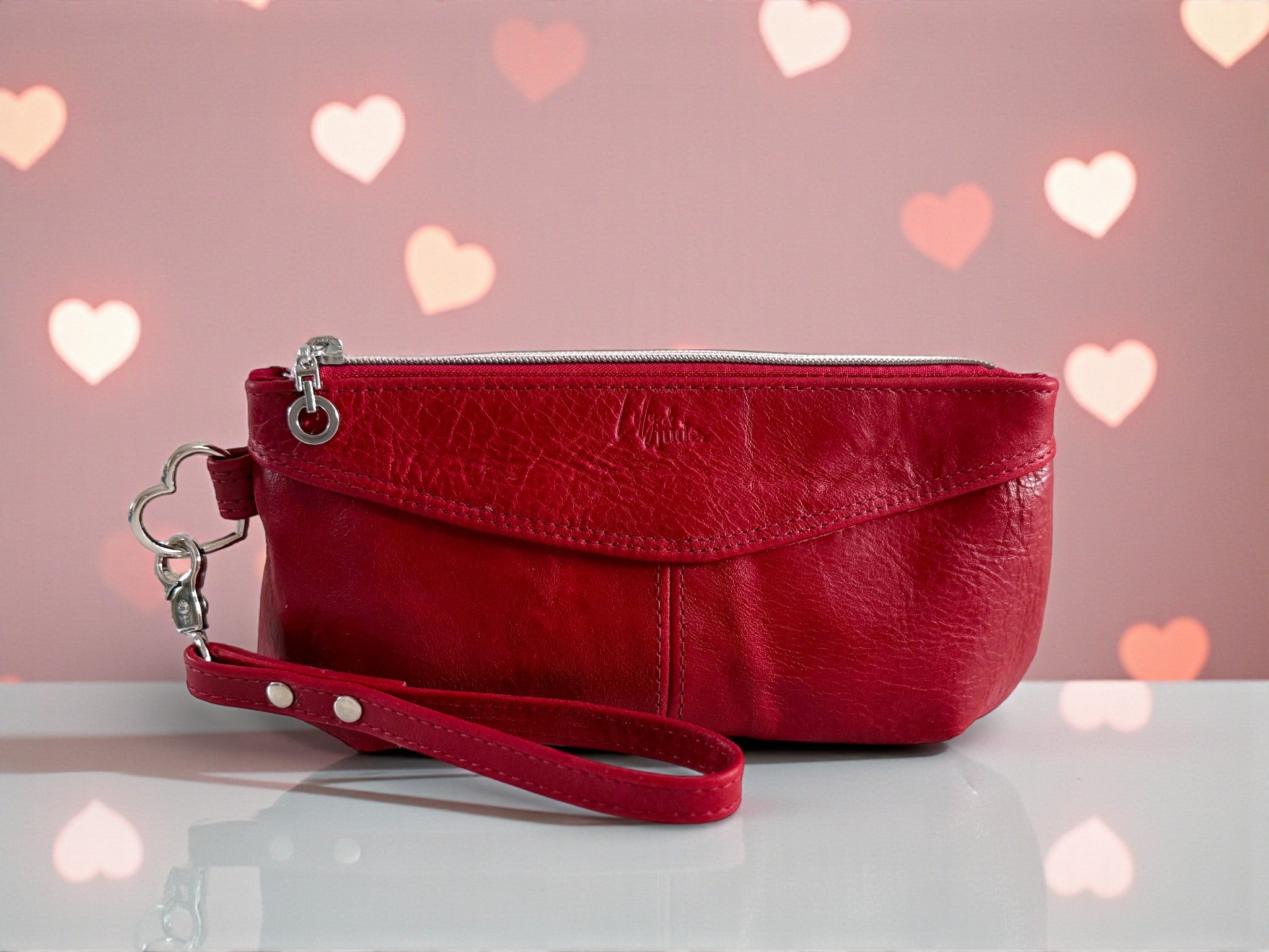 Red leather clutch bag with a wrist strap on a light surface with heart-shaped lights in the background.