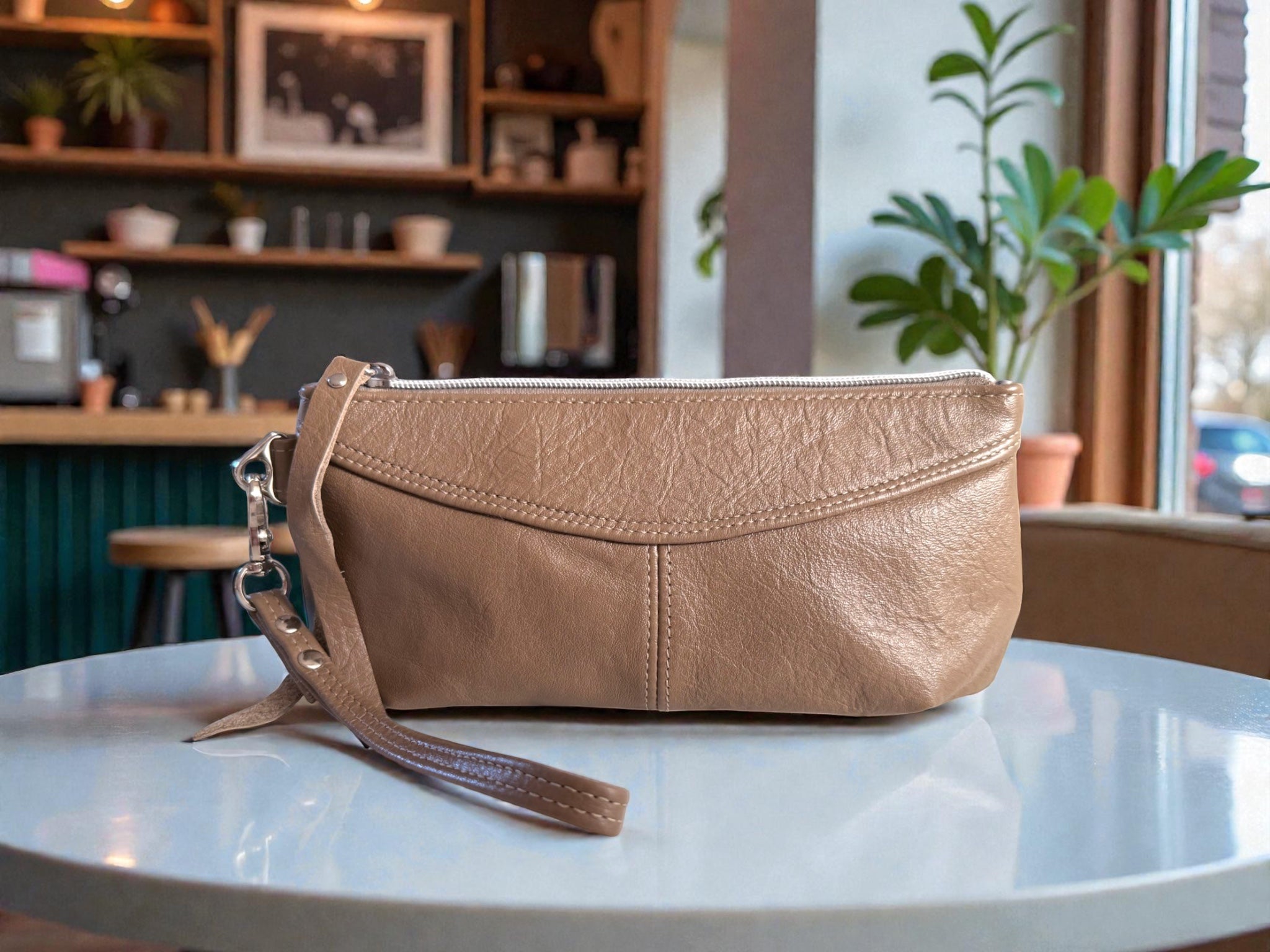 Beige leather clutch, with leather wrist strap on a white table with a blurred cafe background