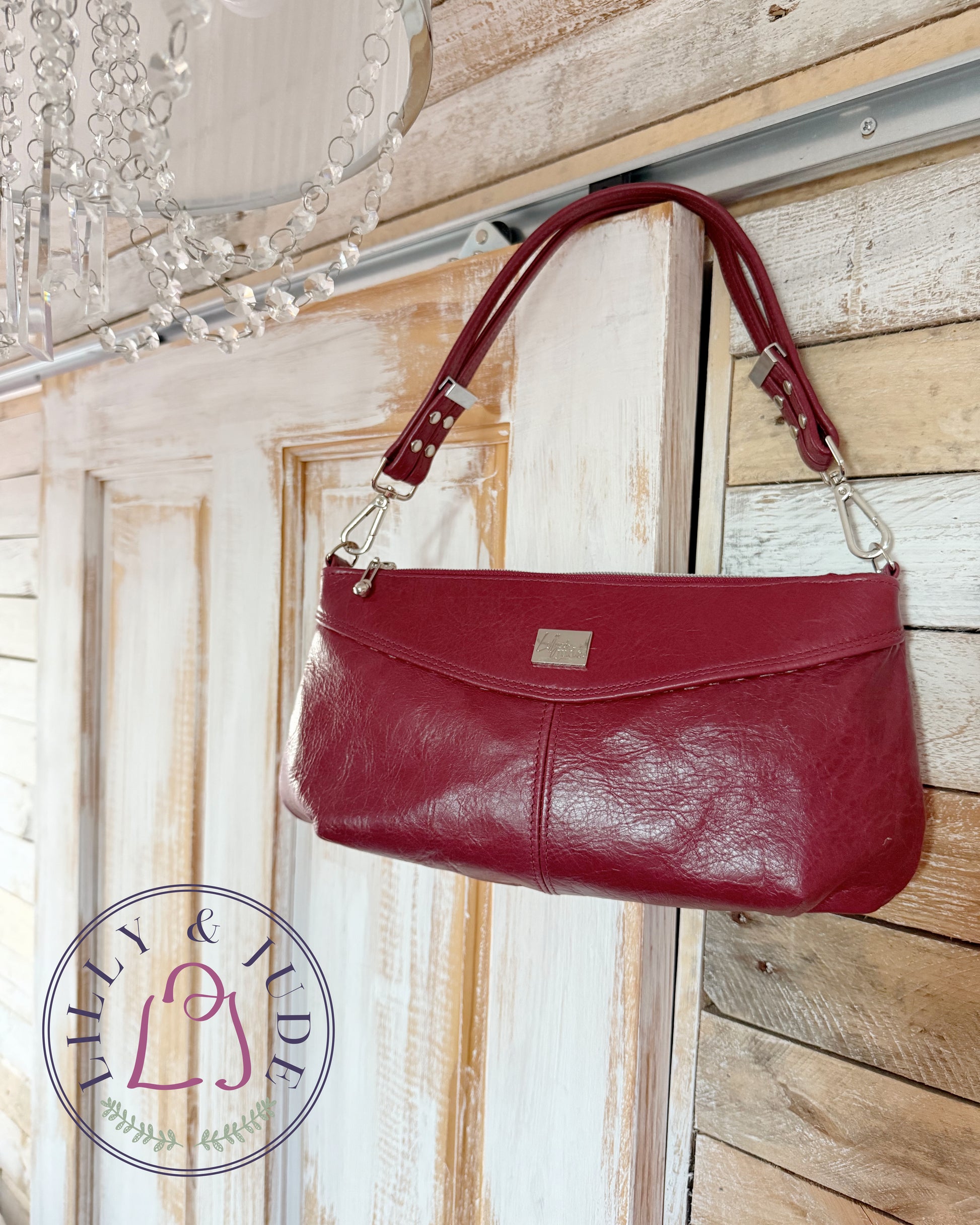 Red handbag hanging on a wooden door with a decorative chandelier in the background. Branded with the Lilly & Jude logo