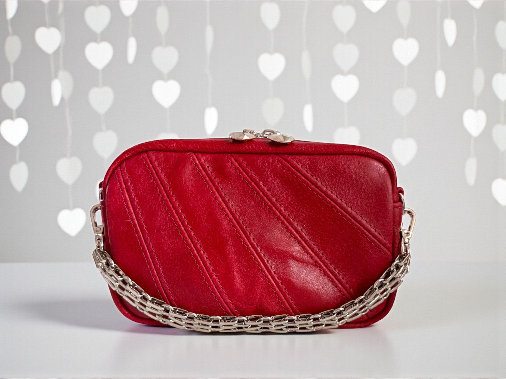 Red leather handbag with a chain detail on a white surface with heart-patterned background