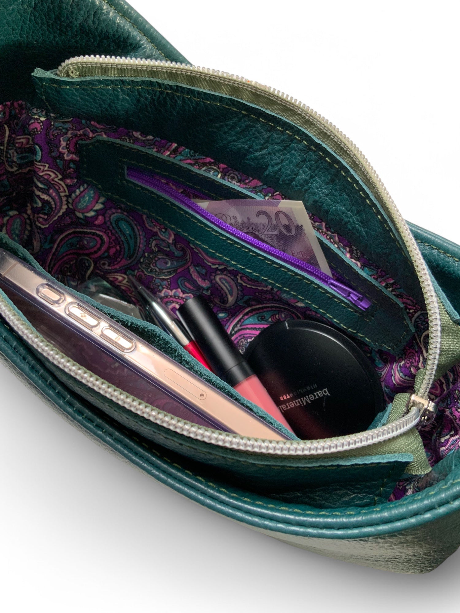 Open teal leather handbag with purple paisley patterned lining and contents on a white background