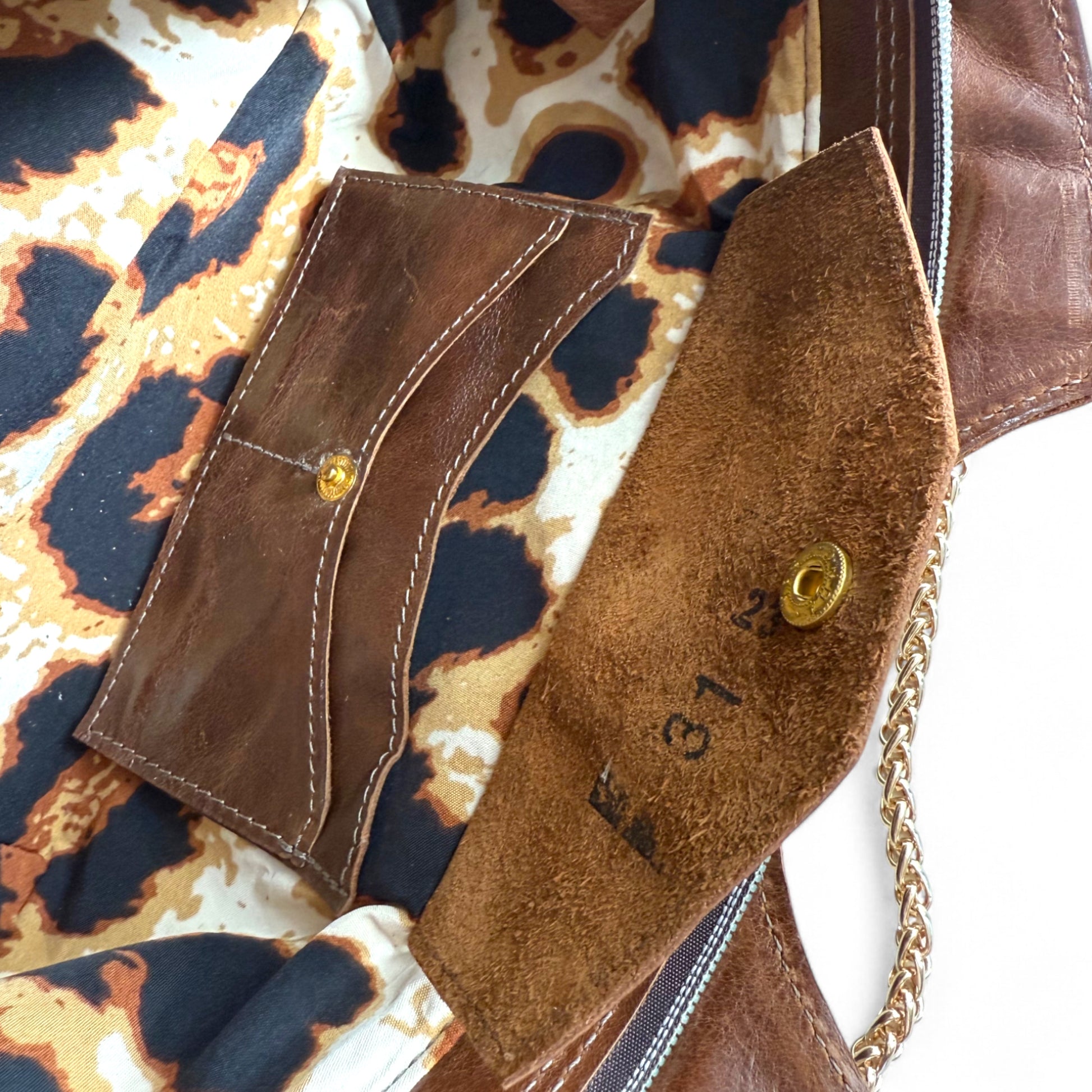 Close-up of a brown leather handbag with a chain detail and the leopard print lining. The image highlights the open integrated leather purse, with the flap open and the markings of the leather maker stamped on the suede side