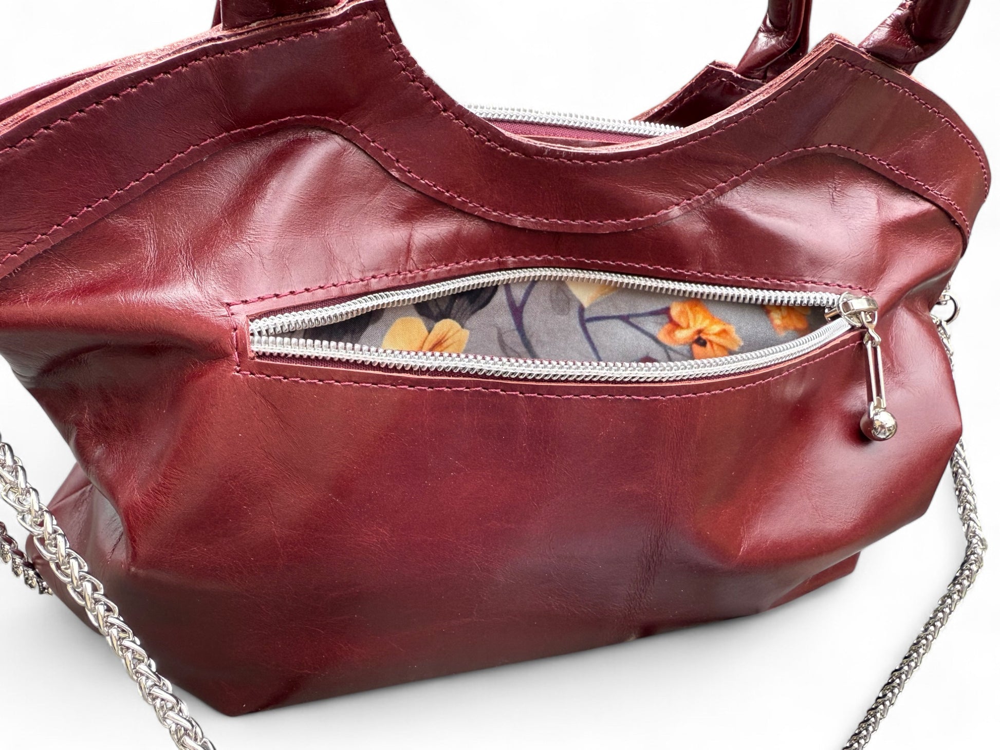 Handcrafted, Bordeaux coloured leather handbag. The exterior zipper pocket is open and is showing the grey contemporary, floral patterned lining.