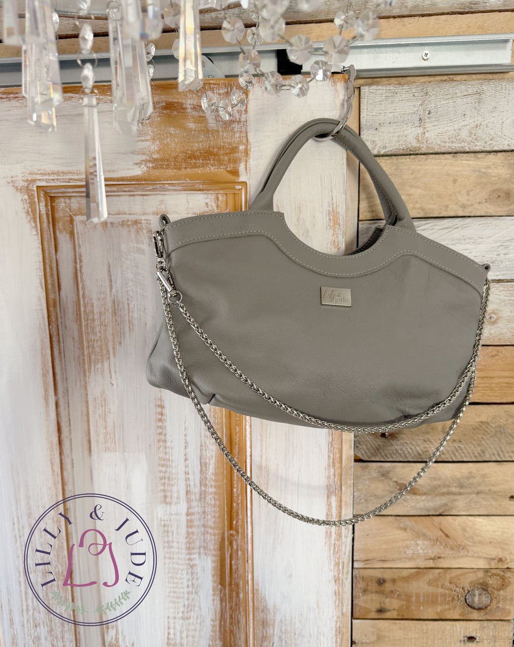 A  grey leather hobo bag with a silver chain strap, hanging against a rustic wood door with a chandelier visible at the top.