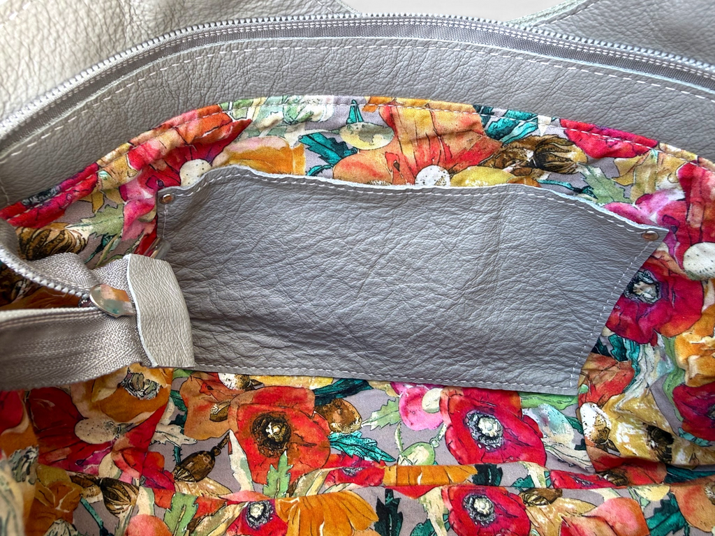 Close-up of the inside of a handbag or tote, showing a colorful floral print lining with red, pink, orange, yellow, and green flowers, a light grey leather pocket, and a matching gray zipper.
