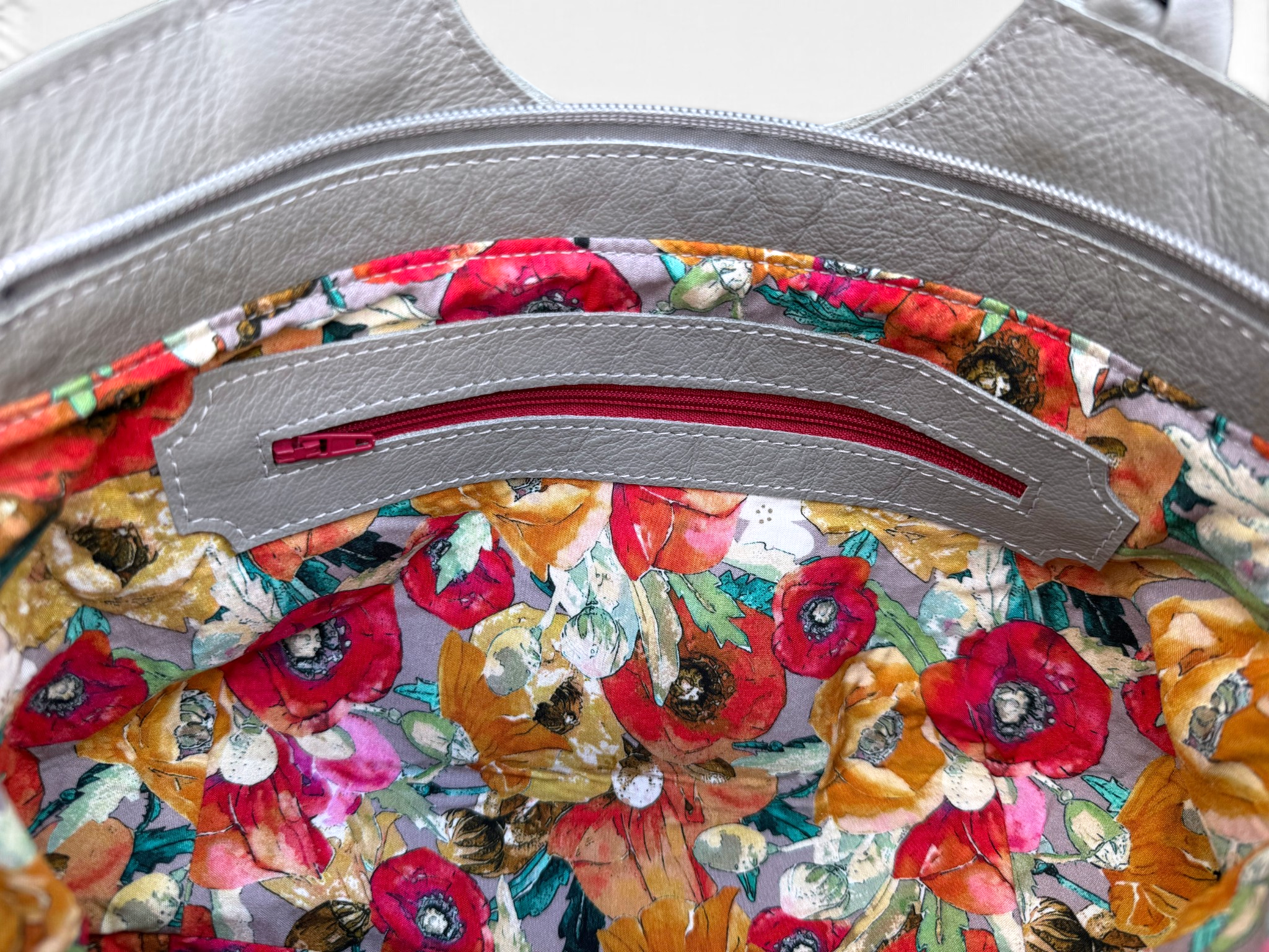 Close-up of the interior of a handbag showing a silver-tone metal zipper track over a colorful floral print lining with red poppy flowers and orange and green leaves.