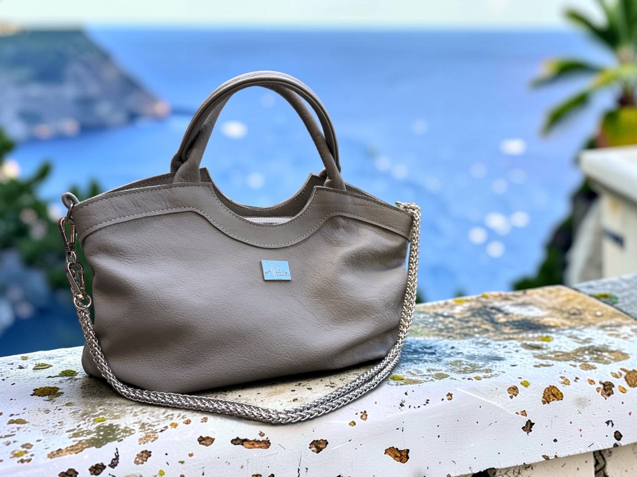 A grey leather slouchy handbag with a silver chain strap, resting on a weathered white stone ledge with a blurred blue sea and green foliage background.