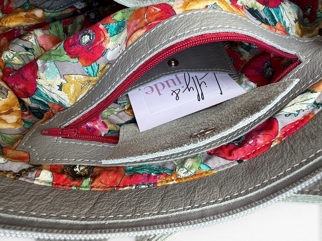 Close-up of a handbag interior showing a grey leather pocket with red zipper trim, a Lilly & Jude business card inside