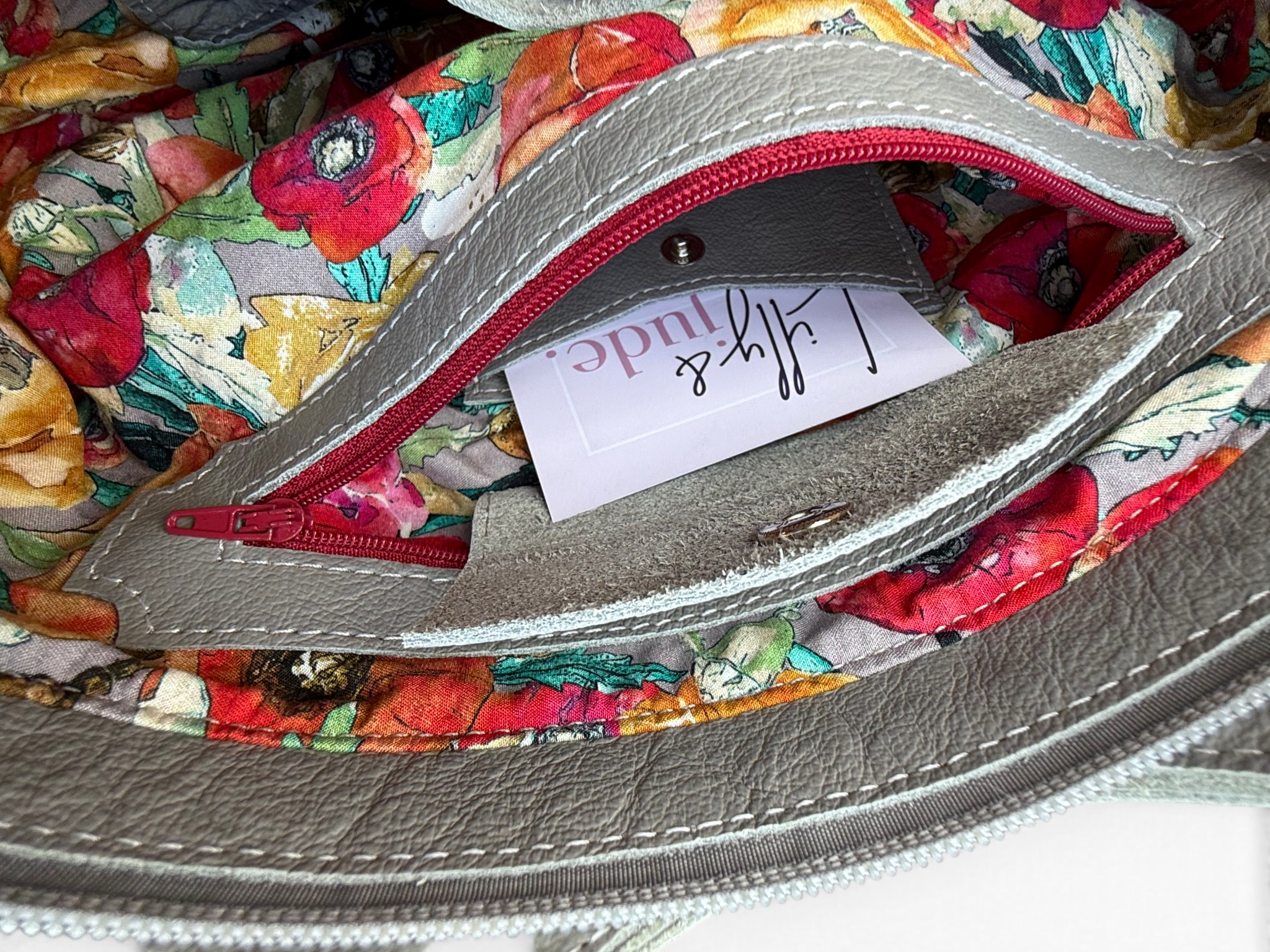 Close-up of a handbag interior showing a grey leather pocket with red zipper trim, a Lilly & Jude business card inside
